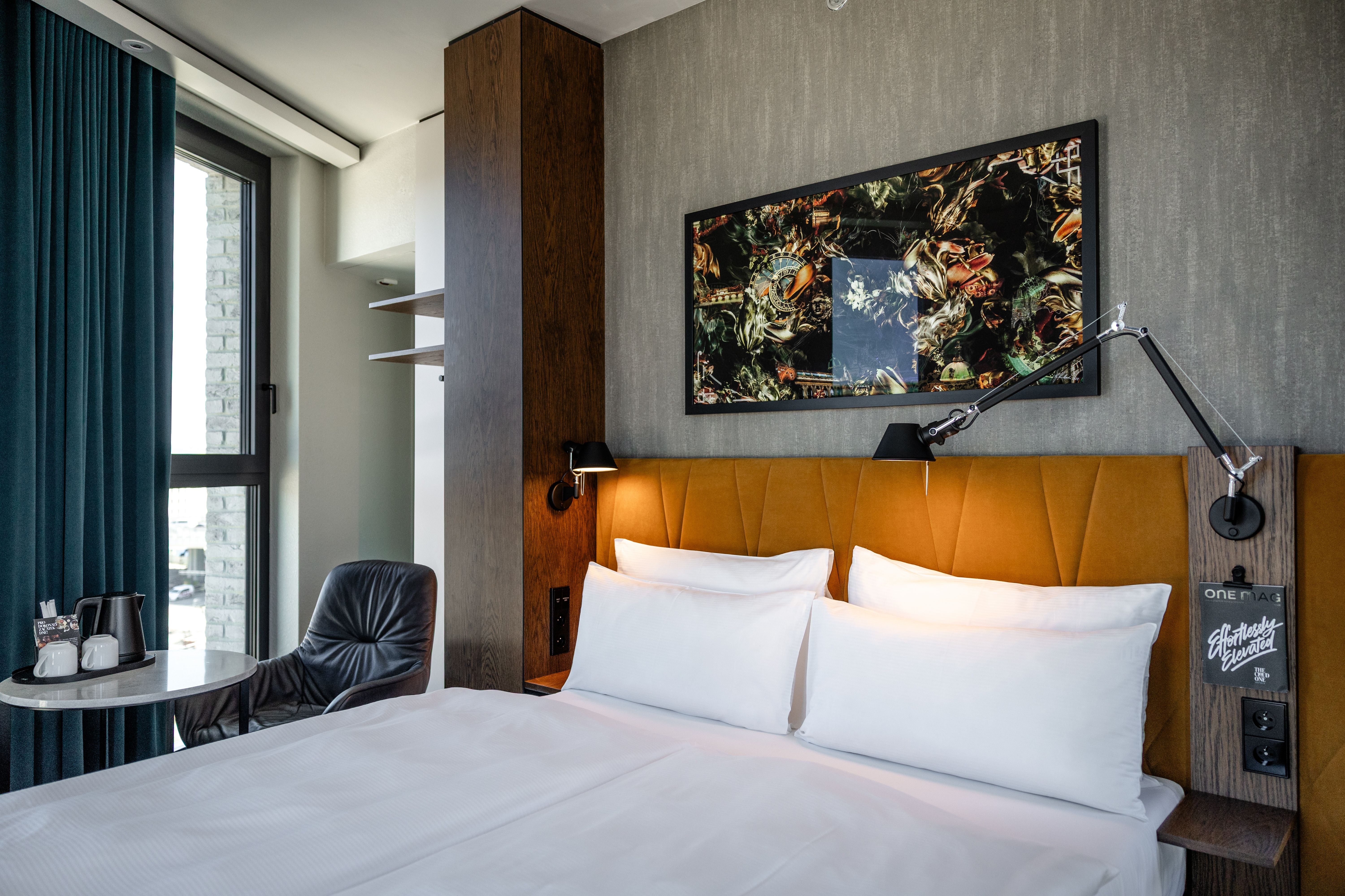 Modern eingerichtetes Hotelzimmer mit komfortablem Doppelbett, warmem Orangenkopfteil, großem Fenster für Tageslicht, moderner Leseleuchte und entspannter Sitzecke mit Fensterblick.