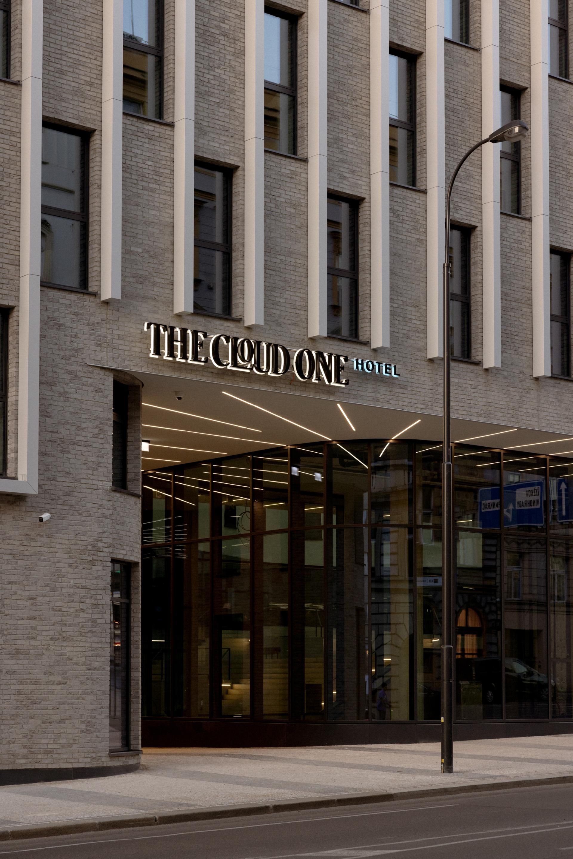 Moderne Hotelfassade aus hellem Stein mit großen Fenstern und beleuchtetem Eingangsbereich des Hotels „The Cloud One“. Urbane Lage an einer ruhigen Straße.