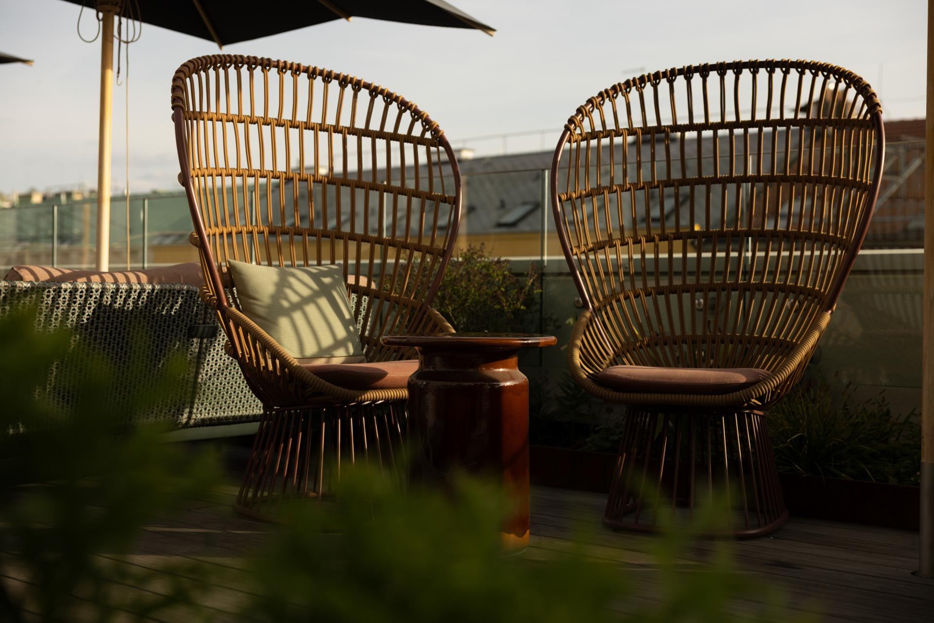 Zwei Rattanstühle mit gemütlichen Kissen stehen auf einer sonnigen Dachterrasse, umgeben von Pflanzen, ideal für entspannte Momente mit Ausblick. Zwei Rattanstühle mit gemütlichen Kissen stehen auf einer sonnigen Dachterrasse, umgeben von Pflanzen, ideal für entspannte Momente mit Ausblick.