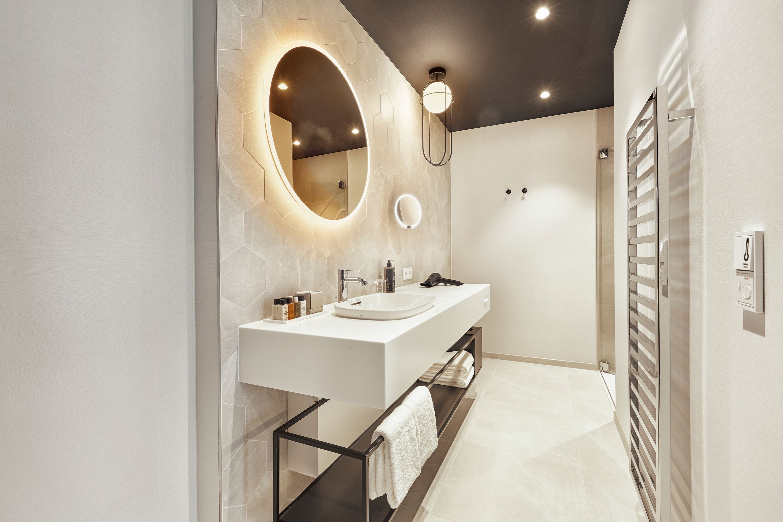 Modernes Badezimmer mit beleuchtetem rundem Spiegel, weißem Waschbecken auf schwarzem Regal und Handtüchern, minimalistischem Design und indirekter Beleuchtung für ein elegantes Ambiente.
