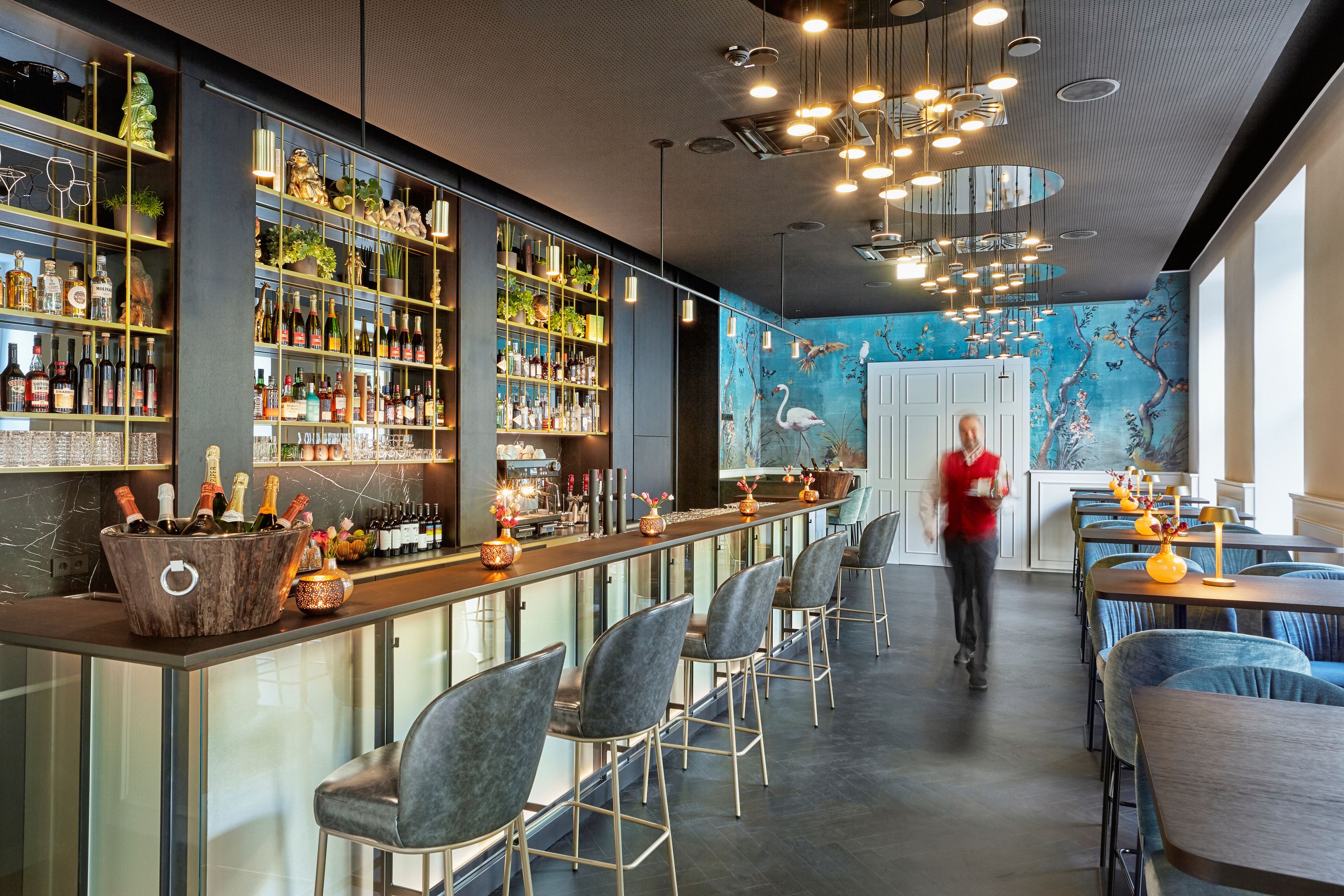 Elegante Hotelbar mit moderner Beleuchtung, bequemen Barhockern und stilvollen Sitzbereichen. Die Wand zeigt ein blaues, florales Wandbild mit Flamingos. Atmosphäre lädt zum Entspannen und geselligen Beisammensein ein.