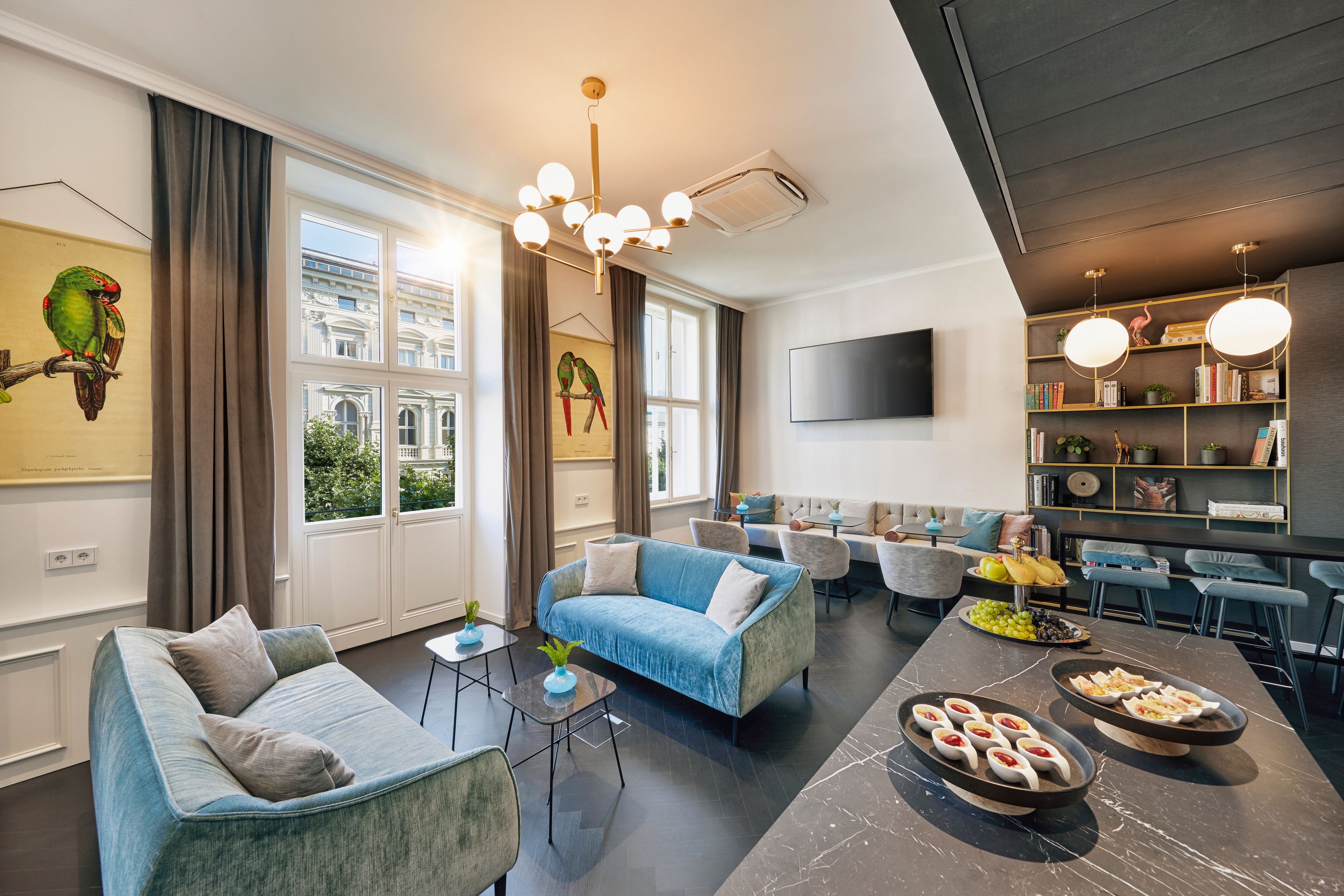 Heller Aufenthaltsraum mit bequemen Sofas, großen Fenstern, stilvoller Beleuchtung und dekorativen Papageienbildern. Bereich zum Entspannen und gemeinschaftlichen Essen mit Snacks und Getränken auf dem Tisch. Gemütliche Atmosphäre für Gäste zum Verweilen und Austausch.