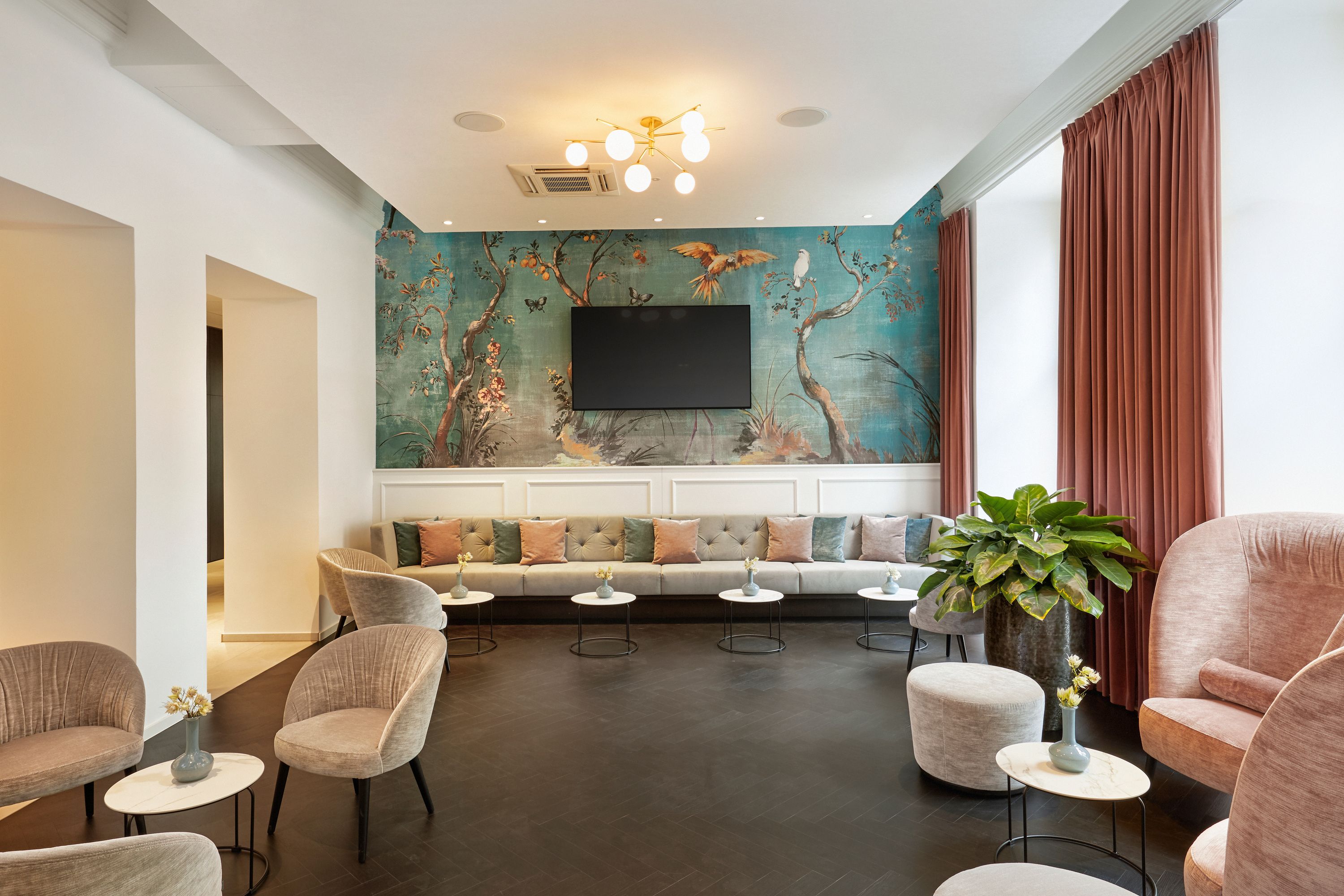 Helles, modernes Lobbyzimmer mit langer, grauer Sitzbank vor einer Wand mit asiatischem Vogel- und Blumenmuster. Große Fenster mit bodenlangen, roten Vorhängen, runde Sessel und kleine Tische schaffen ein einladendes Ambiente zum Entspannen oder Treffen.