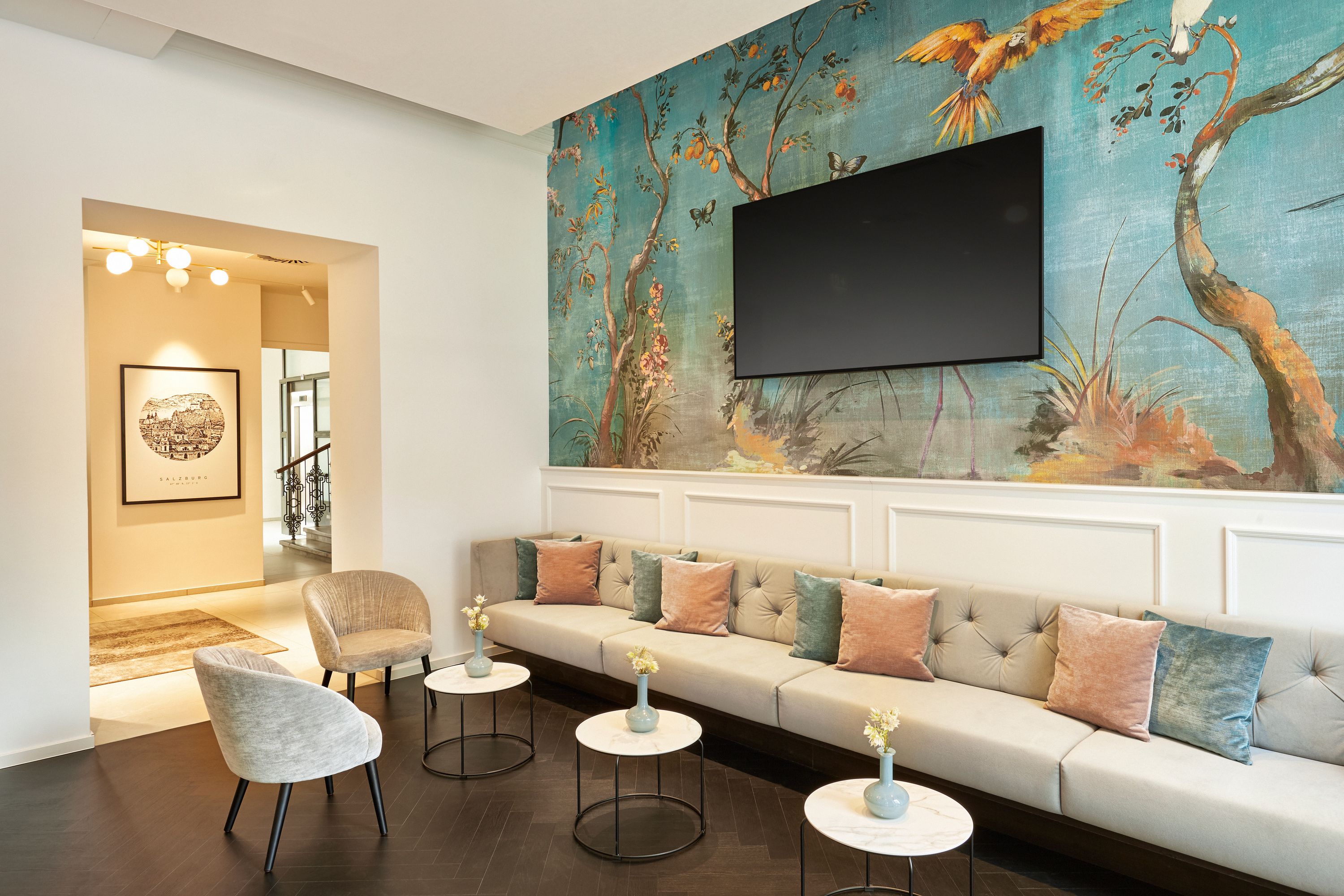 Modern eingerichteter Wohnbereich mit langem Sofa, bunten Kissen, runden Tischen und Vogel-Wandgestaltung, kombiniert mit einem großen Flachbildschirm für entspannte Unterhaltung.