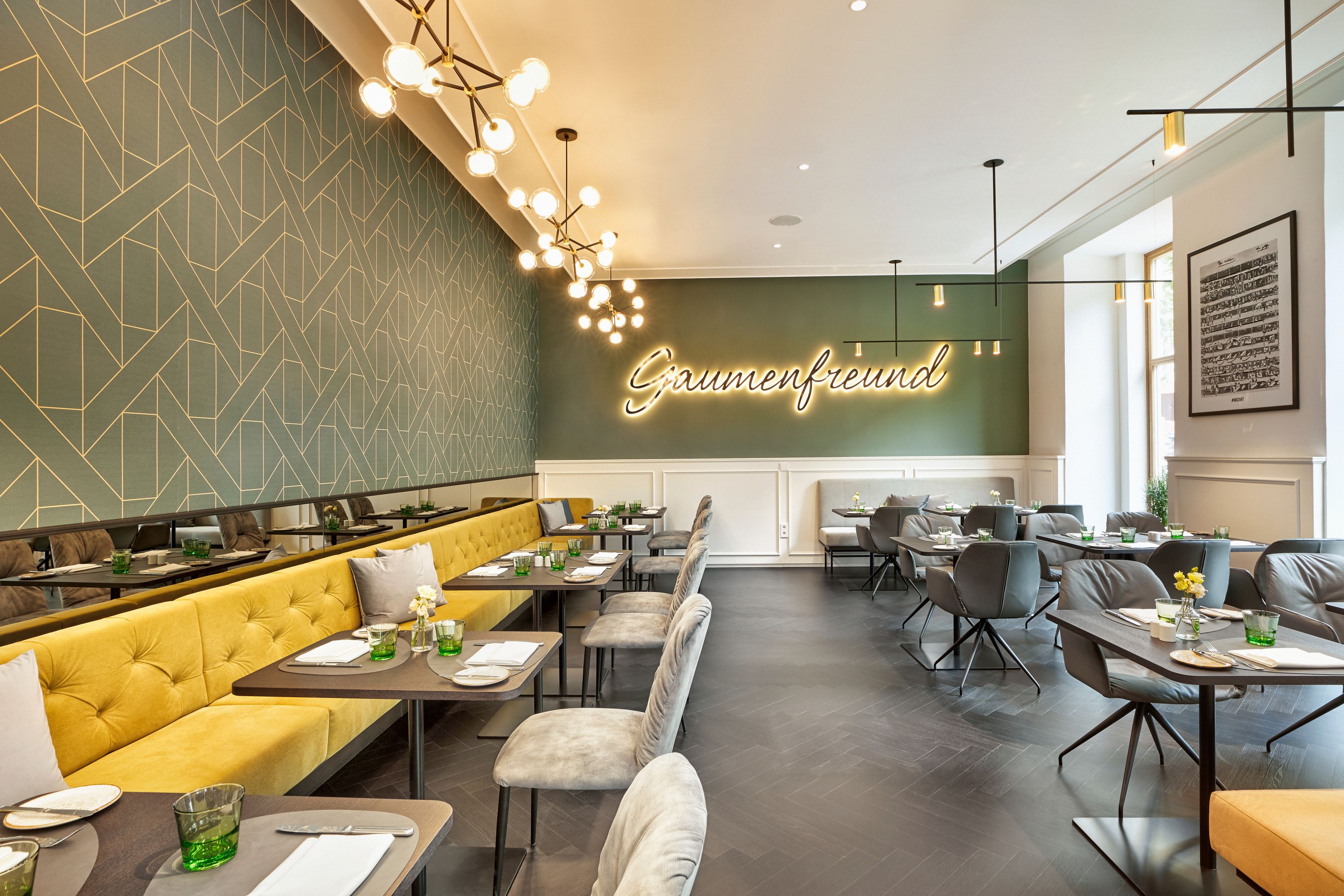 Moderner, gemütlicher Restaurantbereich mit gelber Polsterbank, grauen Stühlen, stilvoller Beleuchtung und einem Leuchtschriftzug „Gaumenfreund“ an der Wand, der einladende Atmosphäre für entspannte Genussmomente schafft.