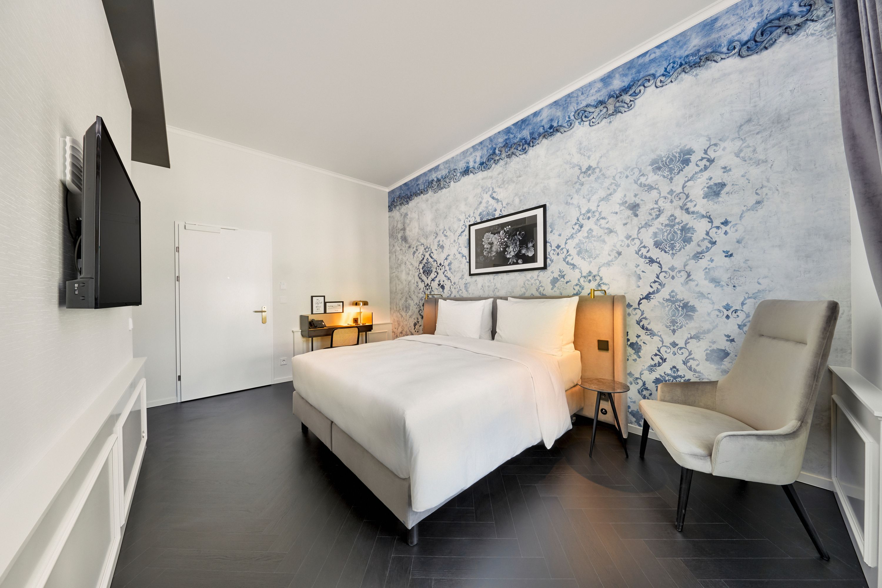 Modern eingerichtetes Hotelzimmer mit großem Doppelbett, stilvoller blauer Wandgestaltung, gemütlichem Sessel und Schreibtisch. Helles, ruhiges Ambiente lädt zum Entspannen und Arbeiten ein.