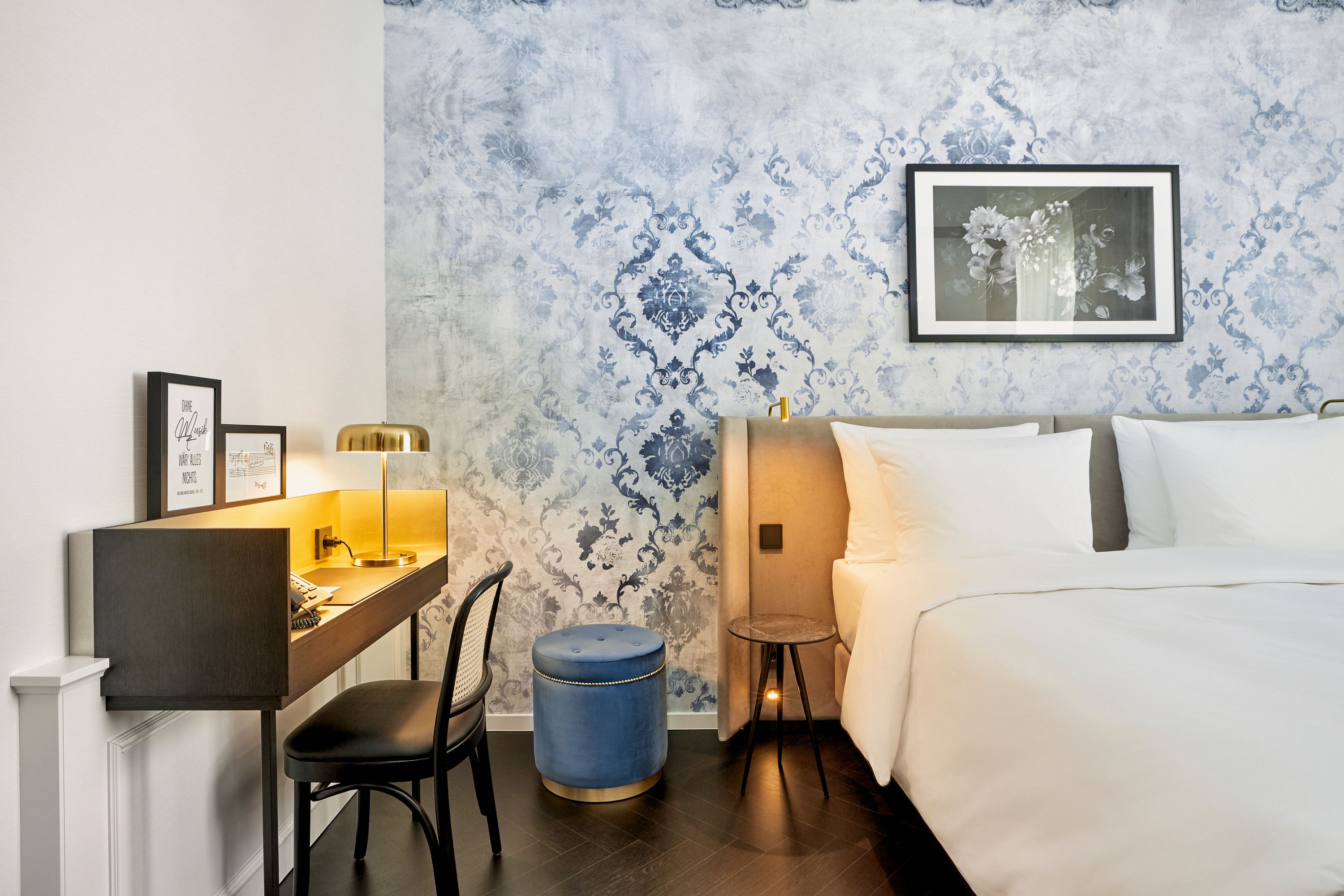 Helles Hotelzimmer mit großem, weiß bezogenem Doppelbett, blauer dekorativer Wand, Schreibtisch mit Stuhl und goldener Schreibtischlampe sowie einem blauen Hocker für zusätzlichen Komfort.