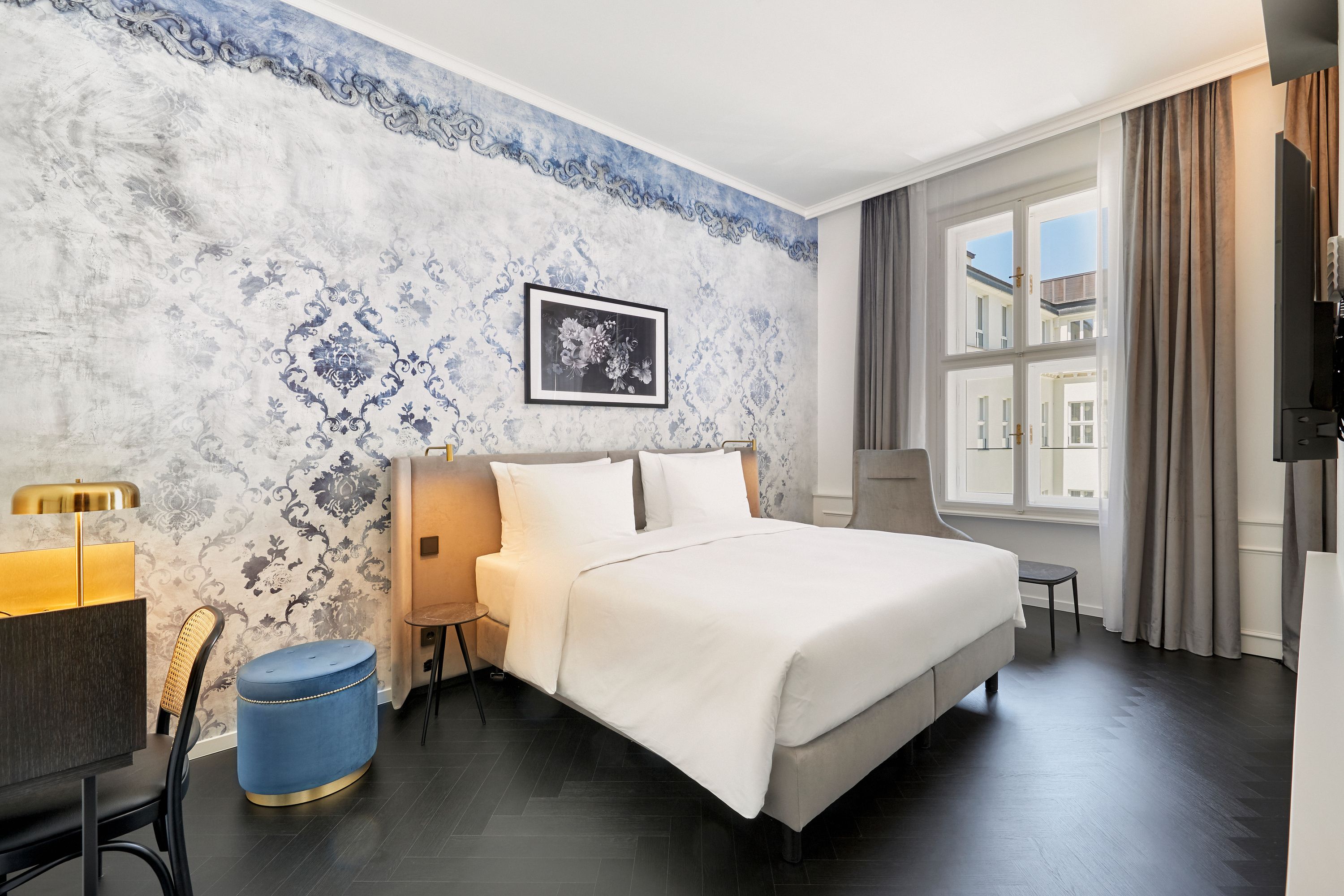Modern eingerichtetes Hotelzimmer mit großem, gemütlichem Doppelbett, stilvoller Wandgestaltung in Blau und Weiß, hellem Tageslicht durch großes Fenster und funktionalem Arbeitsbereich mit Stuhl und Lampe.