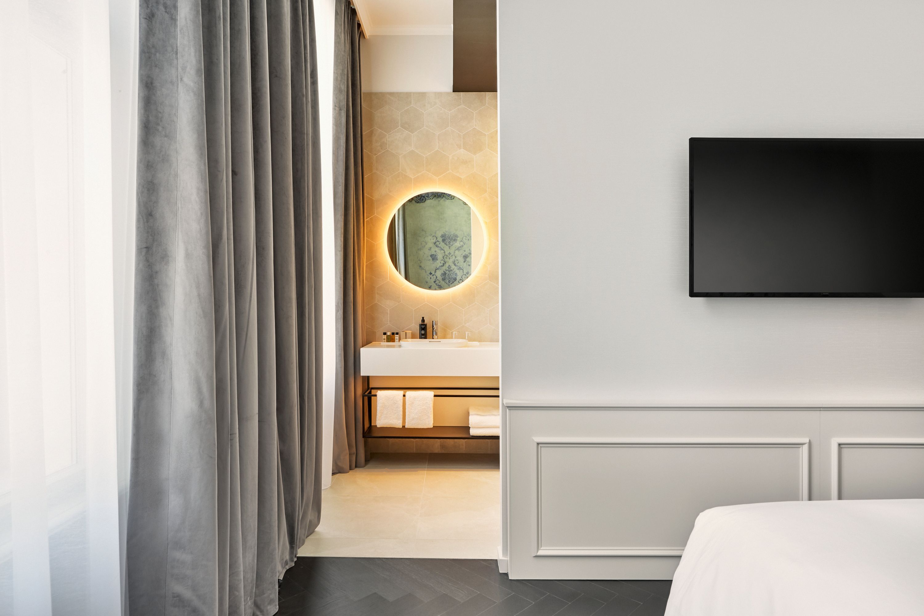Helles Hotelzimmer mit modernem, beleuchtetem Badspiegel, grauen Vorhängen, hellem Waschbeckenbereich und Wand-TV, das eine ruhige und elegante Atmosphäre vermittelt.