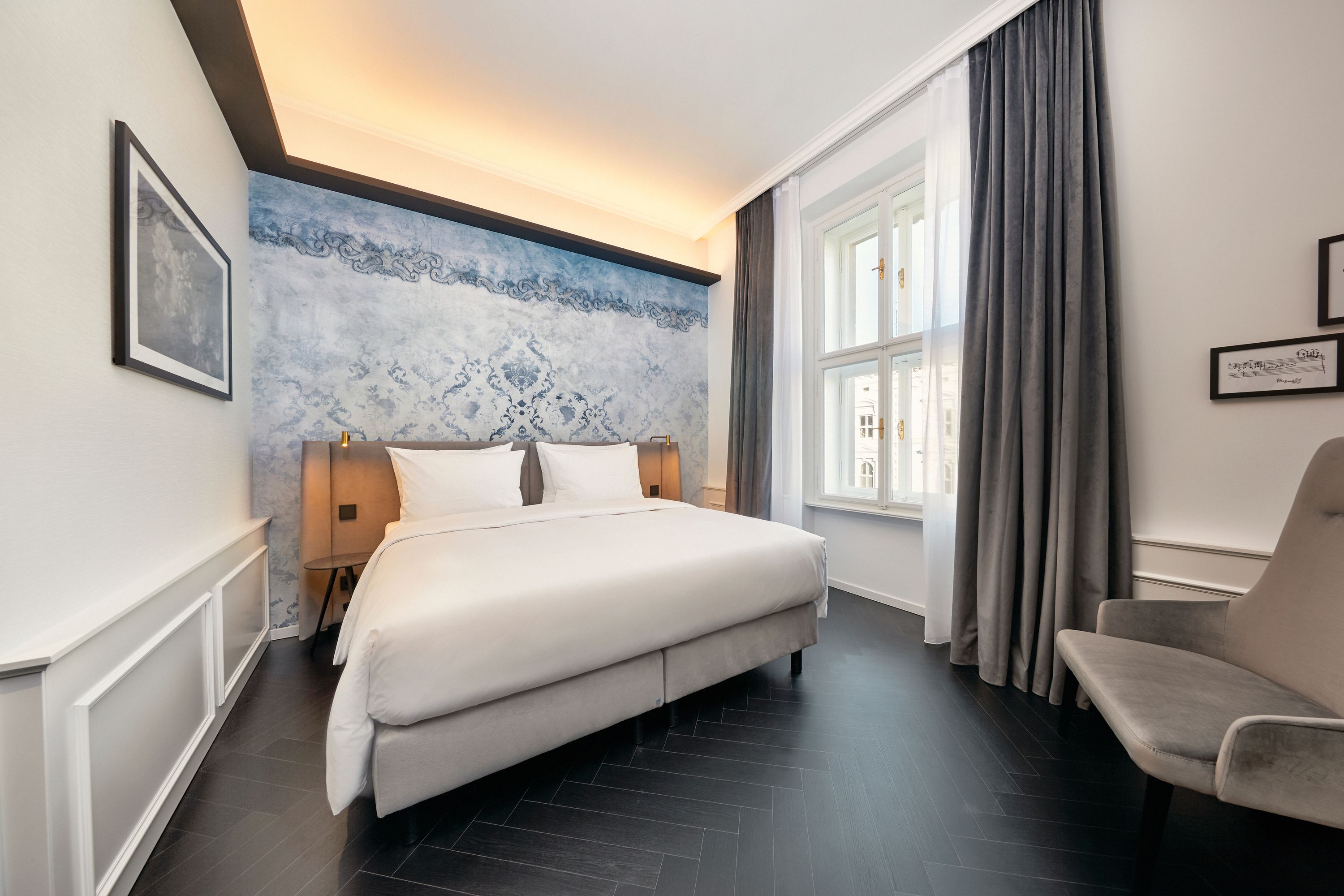 Modernes Hotelzimmer mit großem, bequemem Doppelbett, stilvoller blauer Wandgestaltung, hellem Fenster und Vorhängen, gemütlichem Sessel und dezentem indirektem Licht für eine entspannte Atmosphäre.