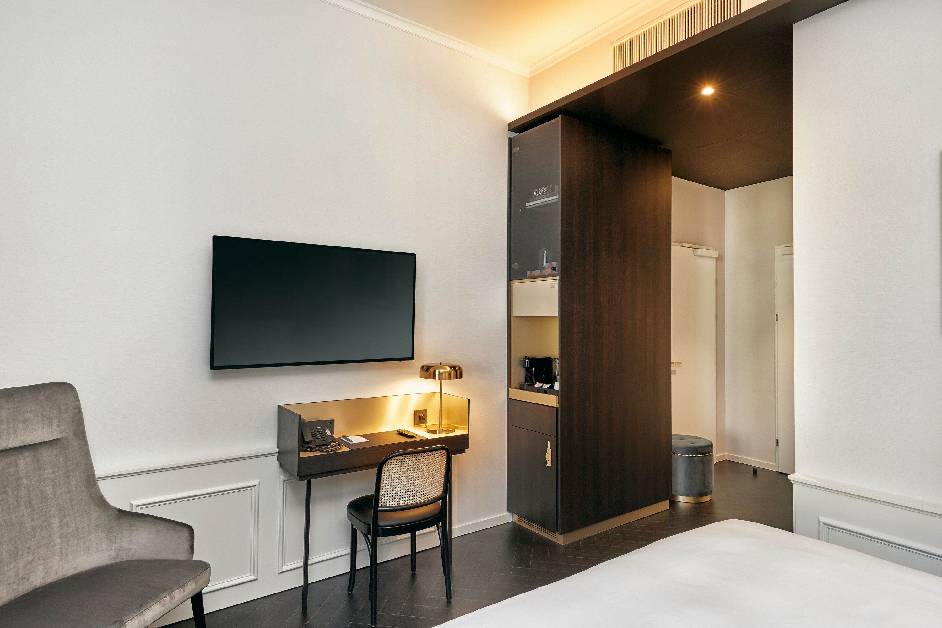 Modern eingerichtetes Hotelzimmer mit Schreibtisch, Stuhl, Flachbildfernseher und gemütlichem Sessel, heller Wandgestaltung und dunklem Holzmöbel, das Stauraum und Zugang zum Flur bietet.