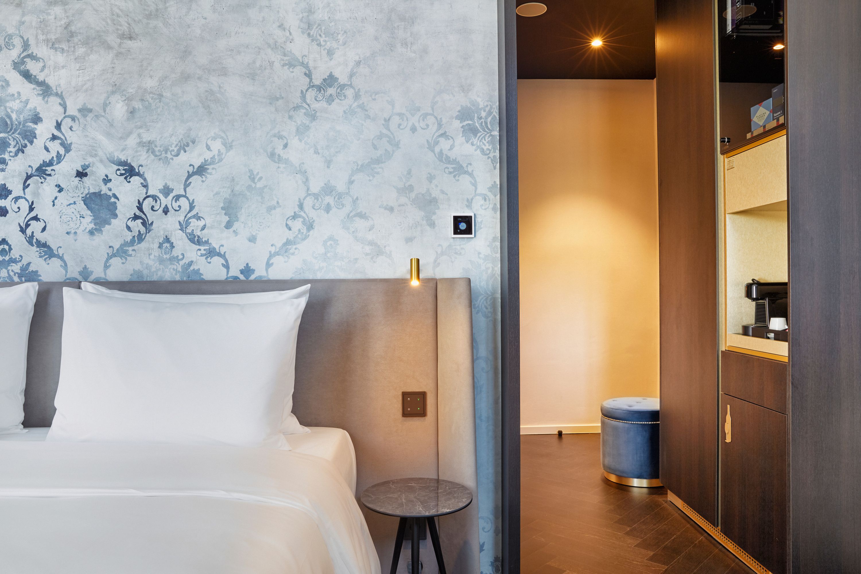 Modern eingerichtetes Hotelzimmer mit gemütlichem Bett, elegantem Kopfteil, sanfter Beleuchtung und stilvollem Beistelltisch. Blick in einen angrenzenden Raum mit warmen Farbtönen und modernen Möbeln. Atmosphäre lädt zum Entspannen und Wohlfühlen ein.
