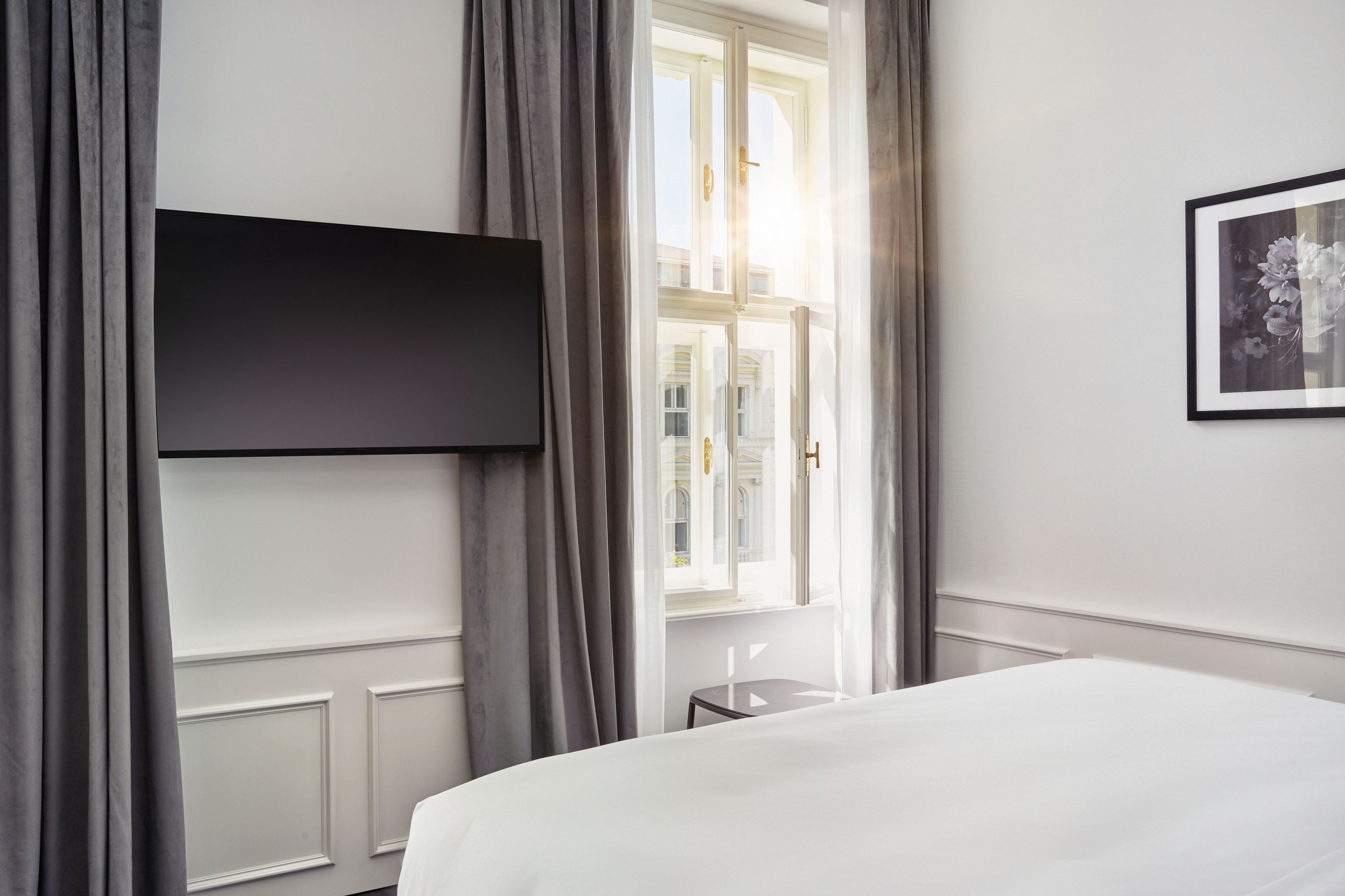 Heller Hotelraum mit offenem Fenster, durch das Sonnenlicht fällt. Weißes Bett, graue Vorhänge und Flachbildfernseher an der Wand vermitteln eine ruhige, moderne Atmosphäre.