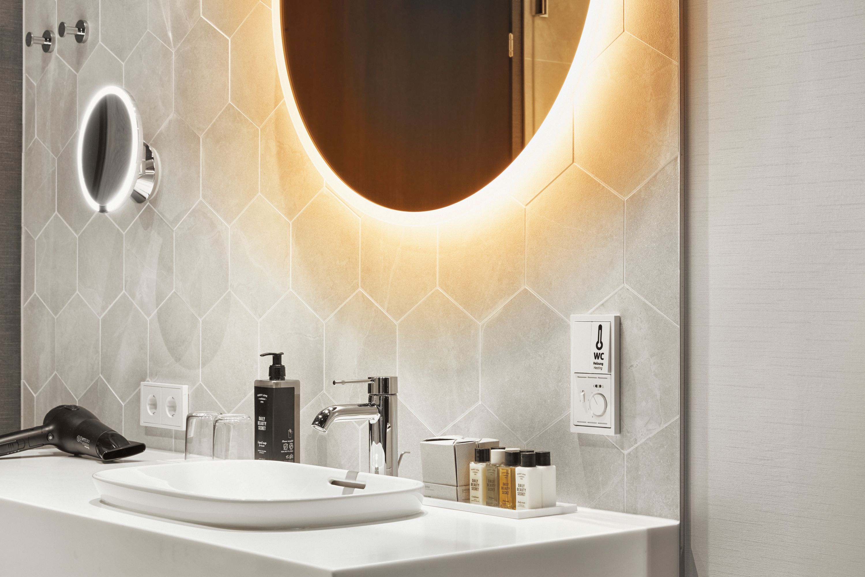 Moderner Hotelbadezimmer-Waschplatz mit beleuchtetem runden Spiegel, weißem Waschbecken, edlen Pflegeprodukten und hellen, hexagonalen Wandfliesen, die eine elegante und einladende Atmosphäre schaffen.