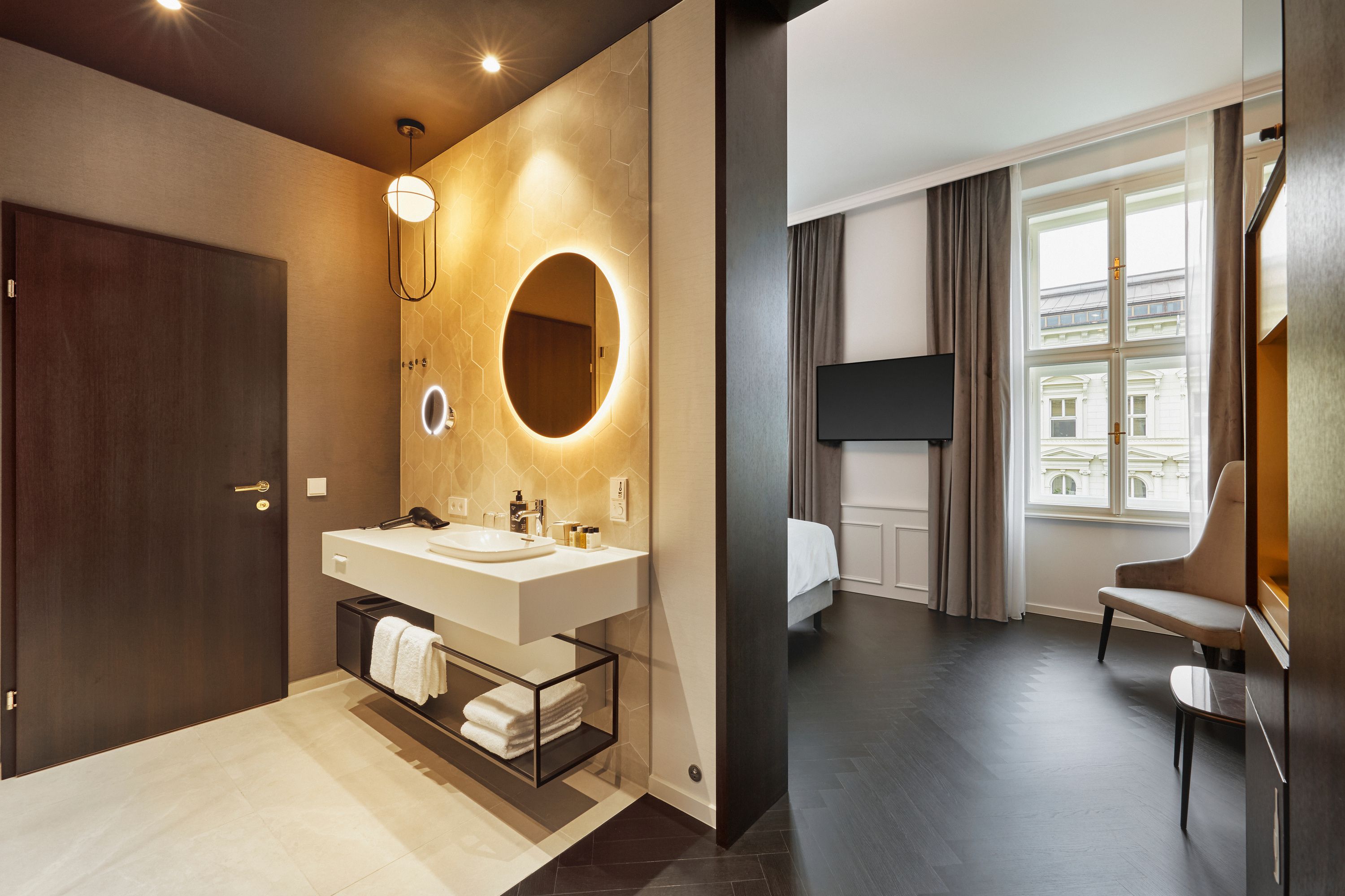 Modernes Hotelzimmer mit hellen, klaren Linien: Helles Badezimmer mit beleuchtetem Rundspiegel und Waschtisch, angrenzendes Schlafzimmer mit großem Fenster, Flachbildfernseher und bequemen Sitzgelegenheiten. Helle und freundliche Atmosphäre.