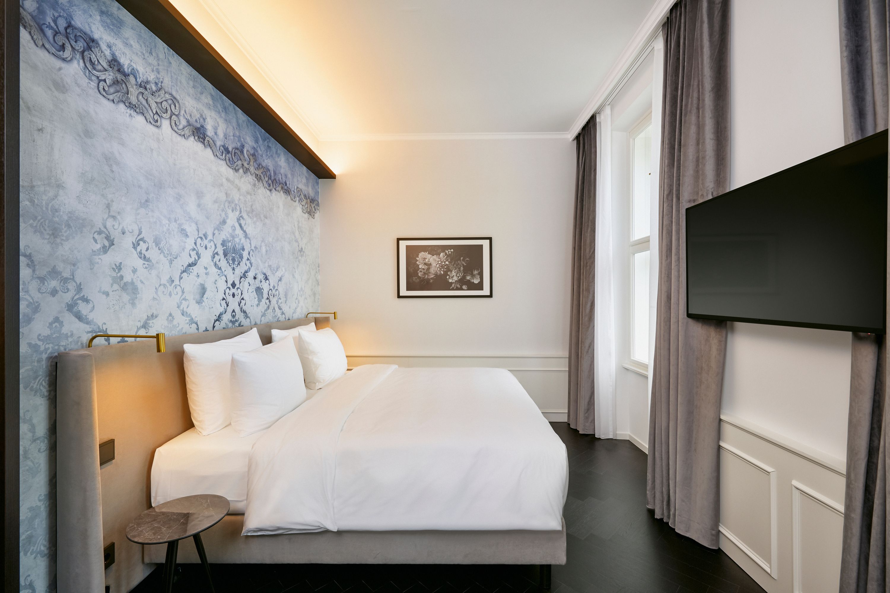 Modernes Hotelzimmer mit großem, weiß bezogenem Doppelbett, stilvoller blauer Wandgestaltung, indirekter Beleuchtung und großem Fenster mit Vorhängen. Flacher Fernseher ist an der Wand montiert. Atmosphärisch ruhig und einladend für erholsamen Aufenthalt.