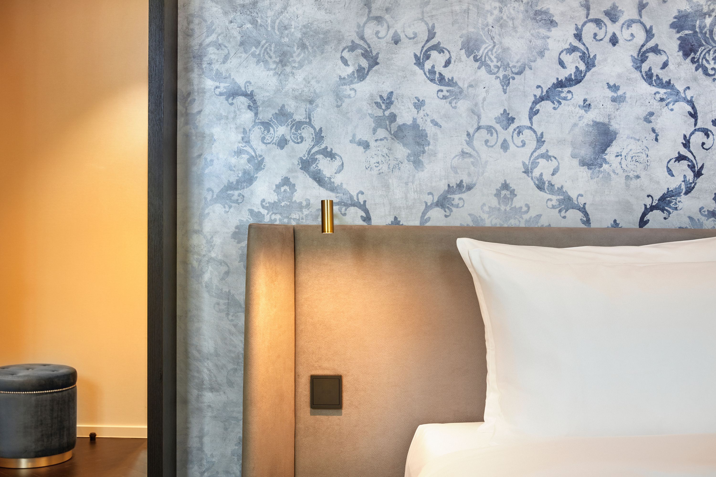 Elegantes Hotelzimmer mit weichem grauem Polsterkopfteil, weißem Bett und stilvoll gemusterter blauer Tapete, mit warmem Licht an der Wand für entspannte Atmosphäre.