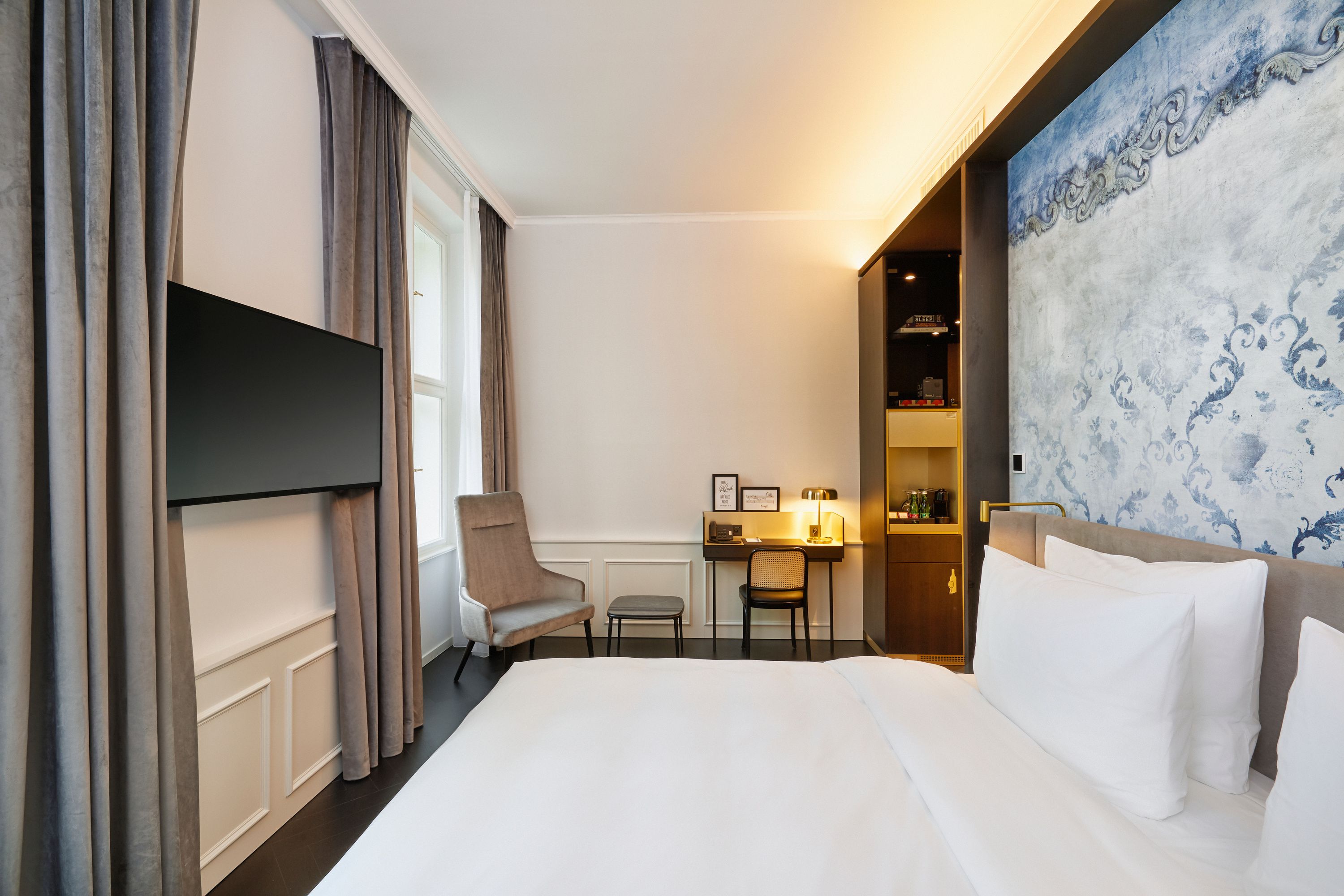 Helles, elegant eingerichtetes Hotelzimmer mit großem, bequemem Doppelbett, stilvoller Wandgestaltung, Schreibtisch mit Lampe und gemütlichem Sessel neben dem Fenster mit Vorhängen, das Raumgefühl wirkt ruhig und einladend für erholsamen Aufenthalt.