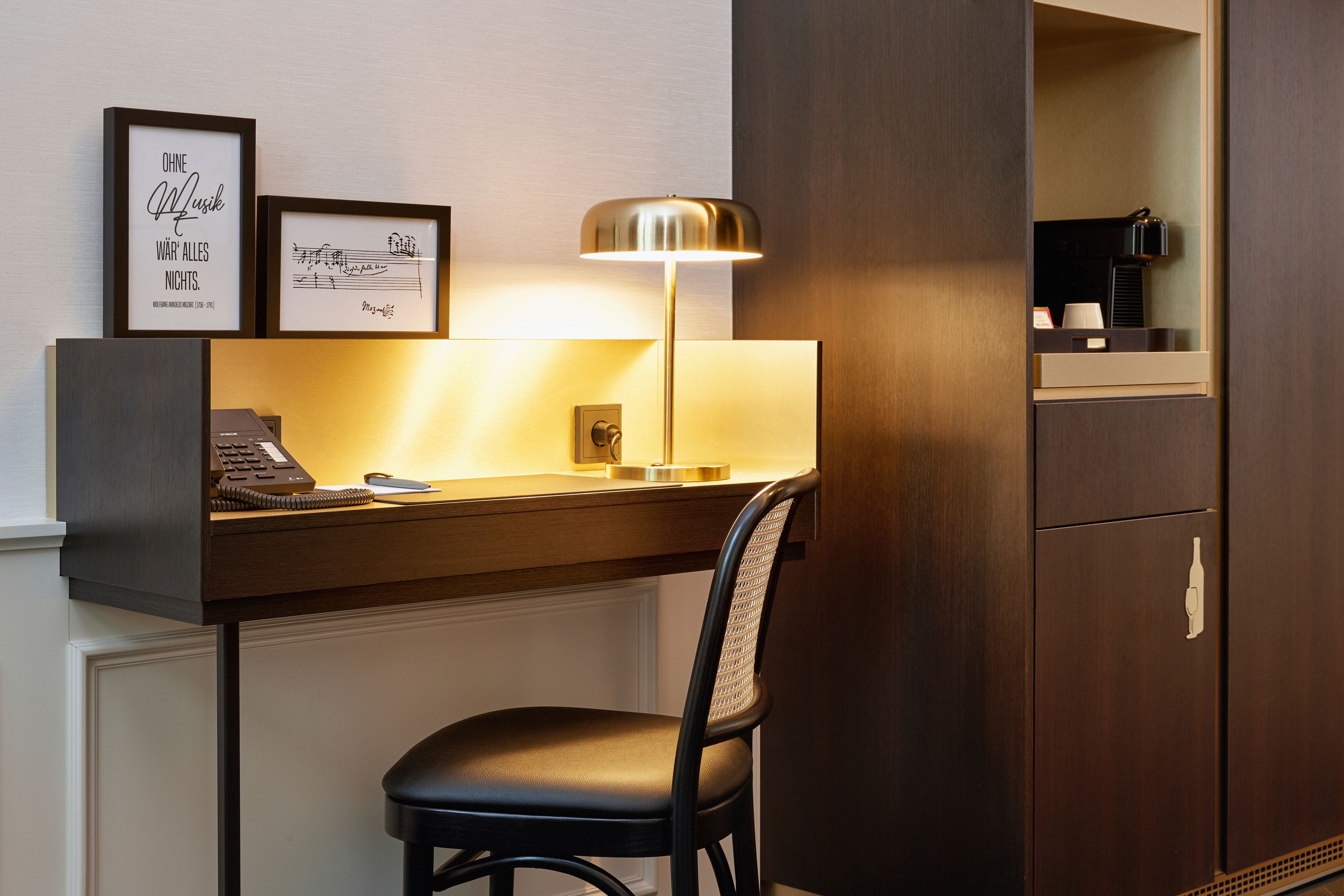Heller Arbeitsbereich mit Schreibtischlampe, Stuhl, Telefon und stilvollen gerahmten Bildern, daneben ein Schrank mit Kaffeezubereiter.