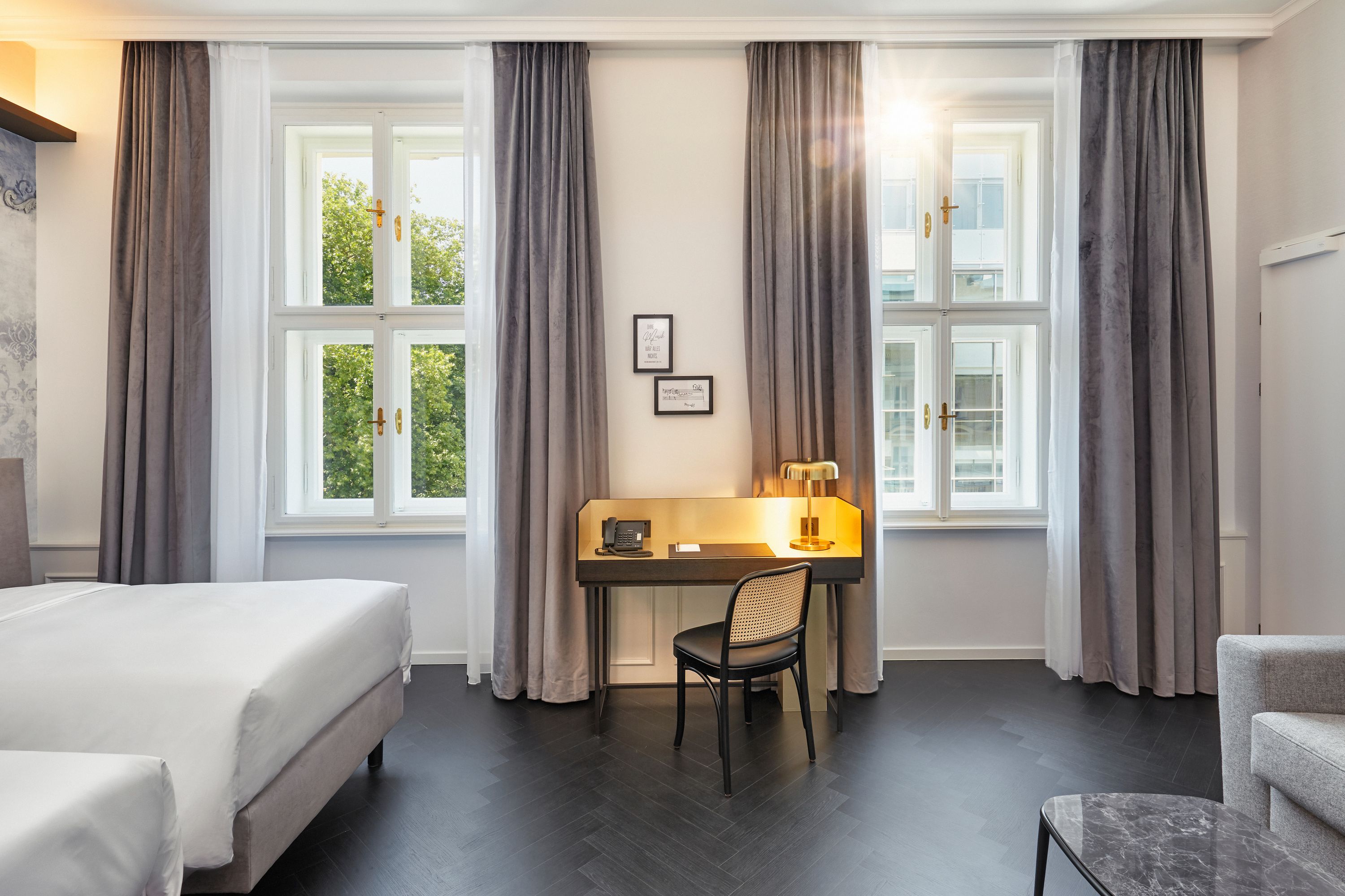 Helles Hotelzimmer mit zwei Betten, Schreibtisch und Stuhl zwischen zwei großen Fenstern, durch die Sonnenlicht hereinfällt. Moderne, schlichte Einrichtung in neutralen Farben vermittelt eine ruhige, einladende Atmosphäre.