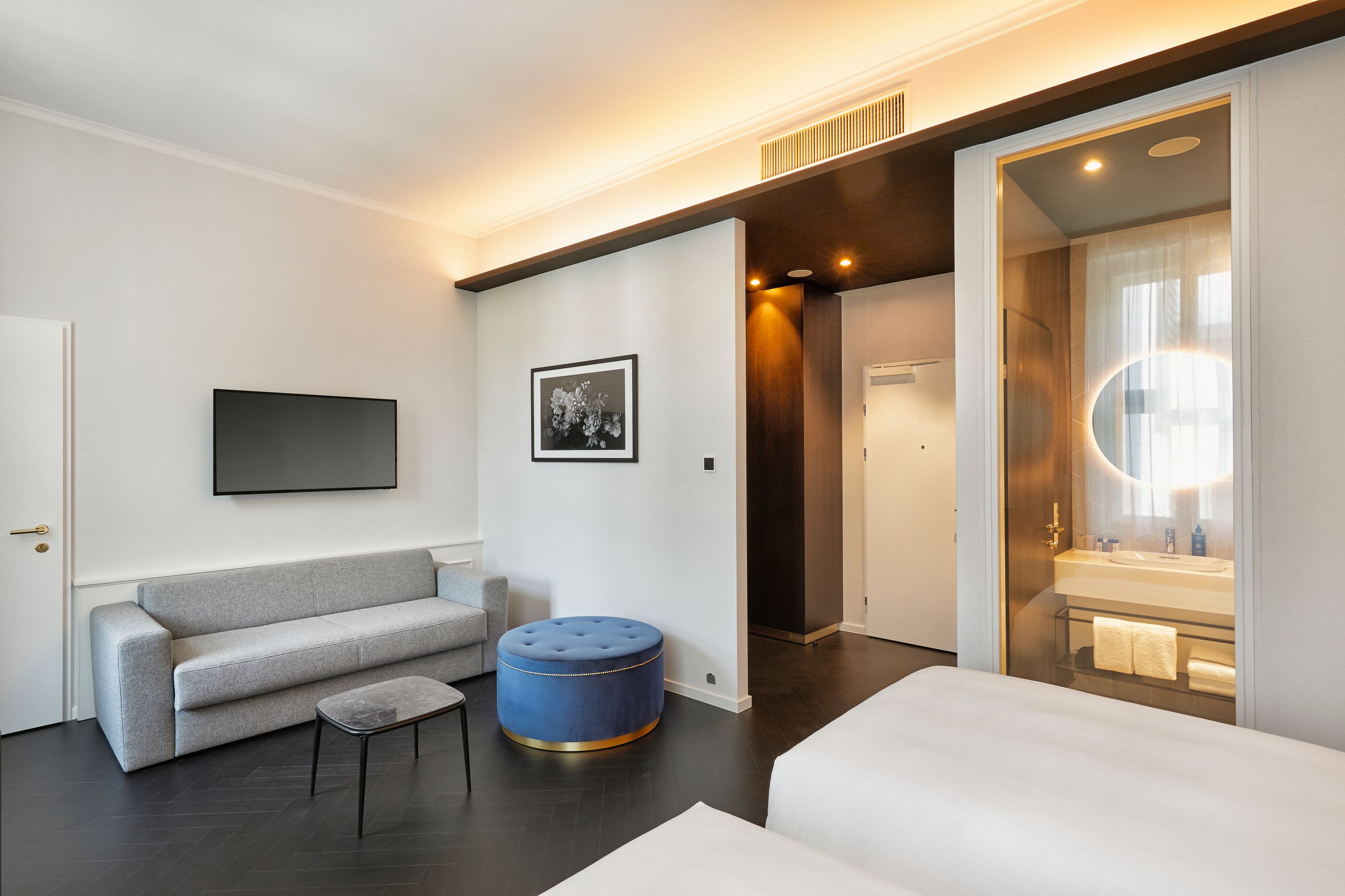 Modern eingerichtetes Hotelzimmer mit zwei Betten, grauem Sofa, blauem Hocker, kleinem Tisch und heller, einladender Atmosphäre. Badezimmer mit beleuchtetem Spiegel sichtbar.
