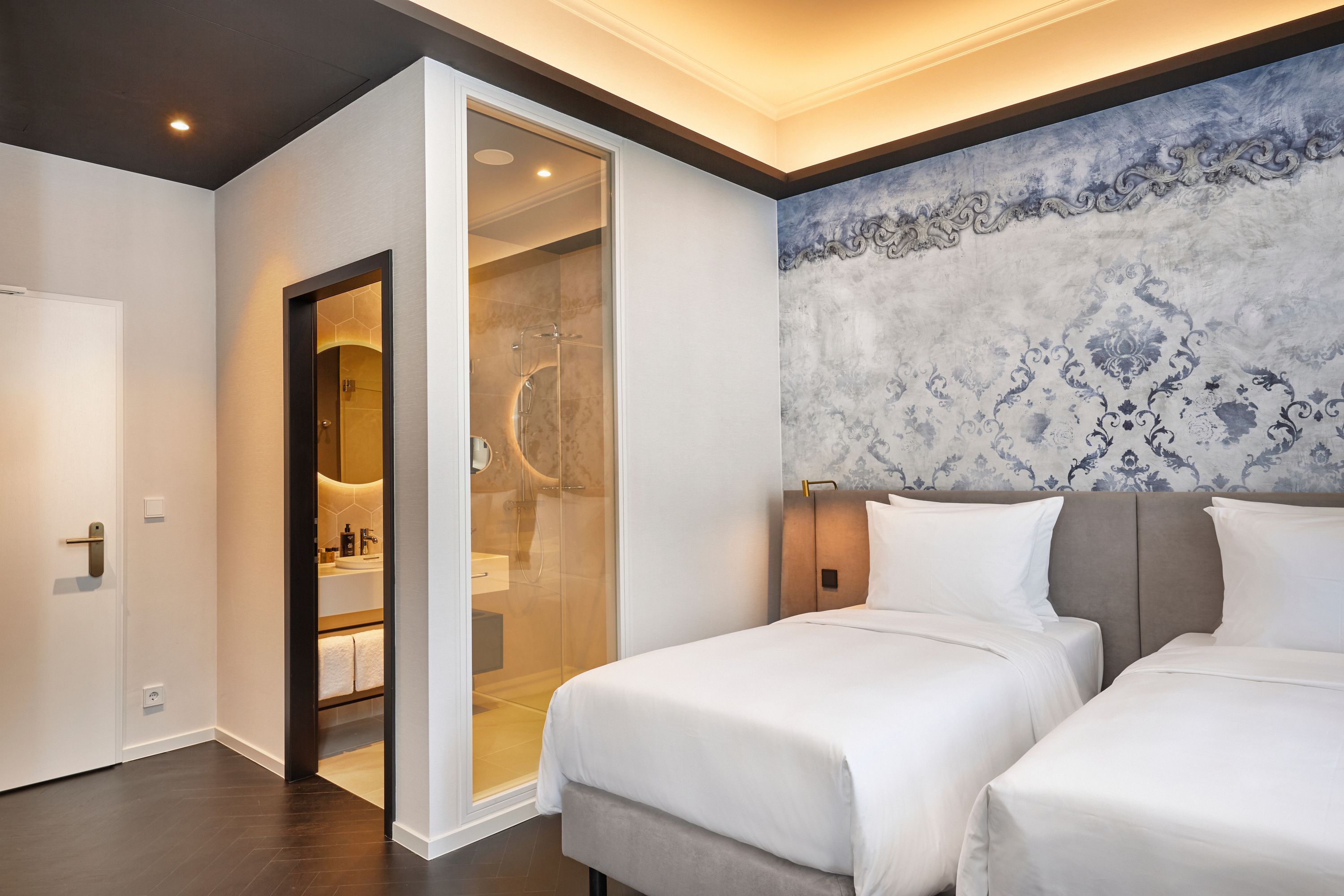Modern eingerichtetes Hotelzimmer mit zwei Einzelbetten, stilvoller Wandgestaltung und indirekter Deckenbeleuchtung. Badezimmer mit Glasabtrennung und moderner Ausstattung sichtbar. Helle, freundliche Atmosphäre für komfortablen Aufenthalt.