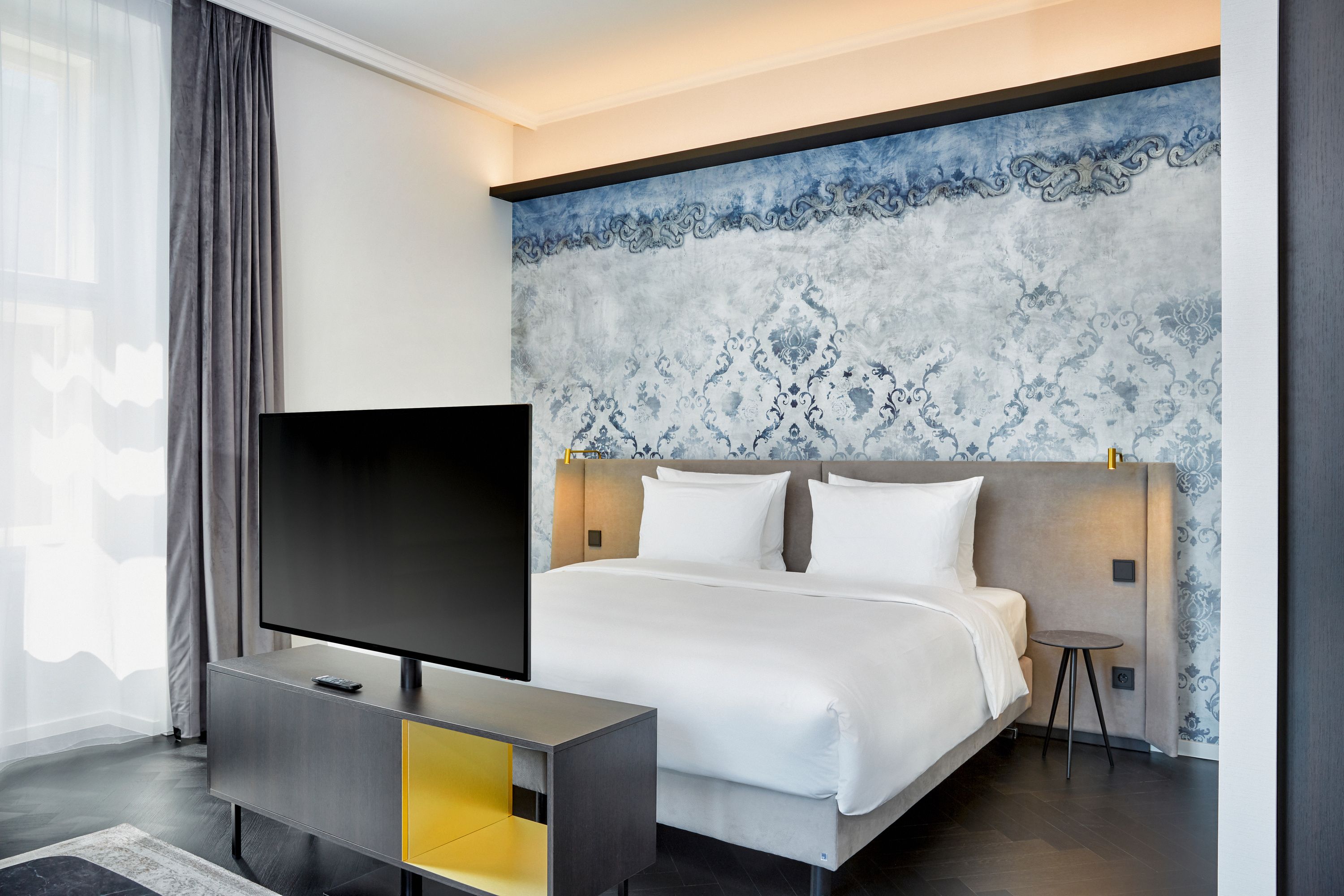 Modern eingerichtetes Hotelzimmer mit großem Doppelbett, dekorativer blauer Wandgestaltung, Fernseher auf einem modernen Lowboard und hellem Tageslicht durch große Fenster mit Vorhängen.