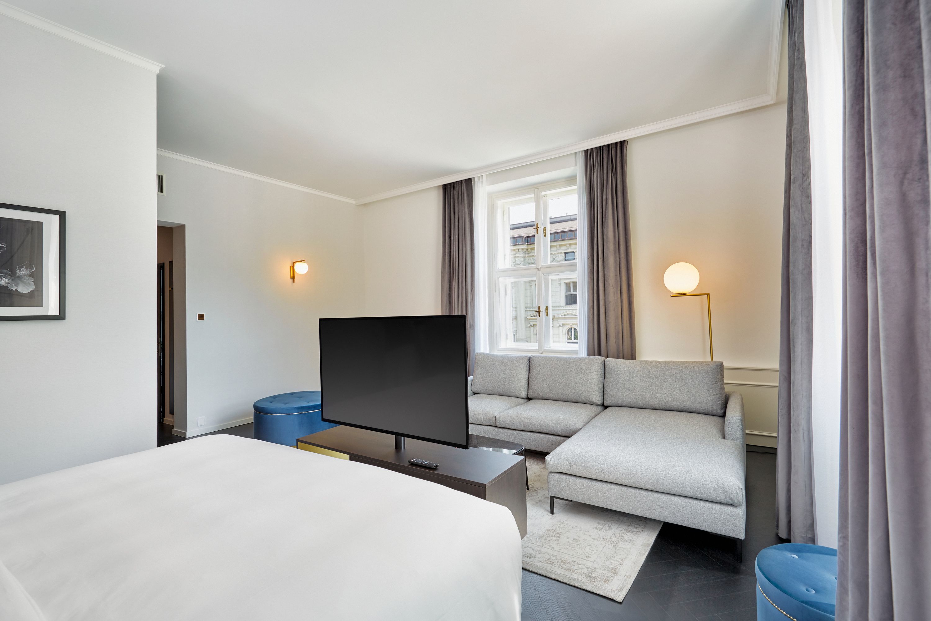 Helles Hotelzimmer mit großem Doppelbett, grauem Ecksofa, Flachbildfernseher und Fenster mit Blick auf Nachbargebäude. Dezente Beleuchtung und schlichte, moderne Einrichtung schaffen eine gemütliche Atmosphäre.