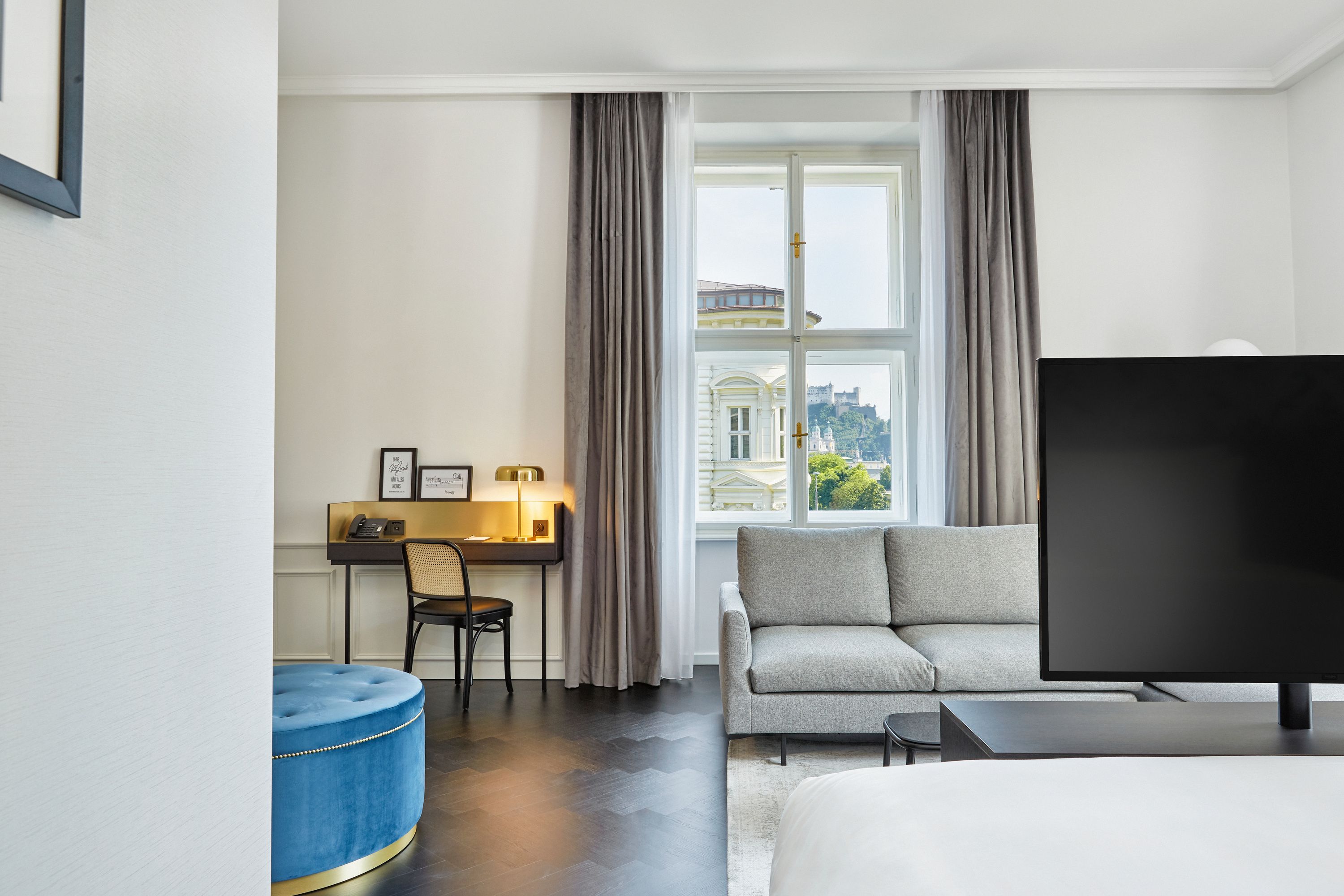 Helles Hotelzimmer mit großem Fenster, das eine Burg und Stadtblick bietet. Komfortable Sitzecke mit grauem Sofa, moderner Schreibtisch mit Lampe und Stuhl, großer Flachbildfernseher, eleganter Holzboden und dezente Farbgestaltung.