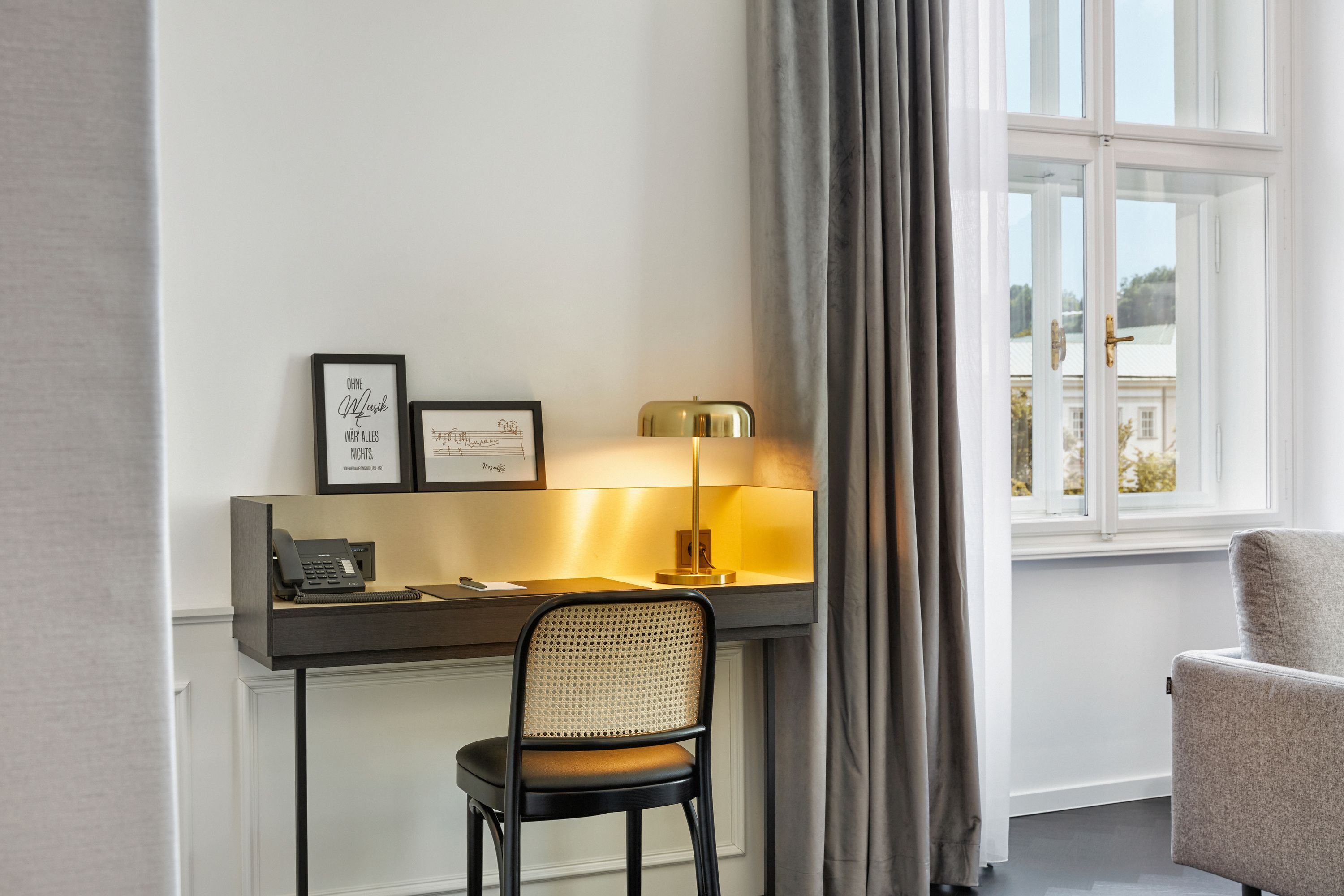 Heller, moderner Arbeitsbereich mit Schreibtisch, Stuhl und Tischlampe neben einem großen Fenster, das Tageslicht hereinlässt. Angenehme Atmosphäre für konzentriertes Arbeiten oder Schreiben im Hotelzimmer.