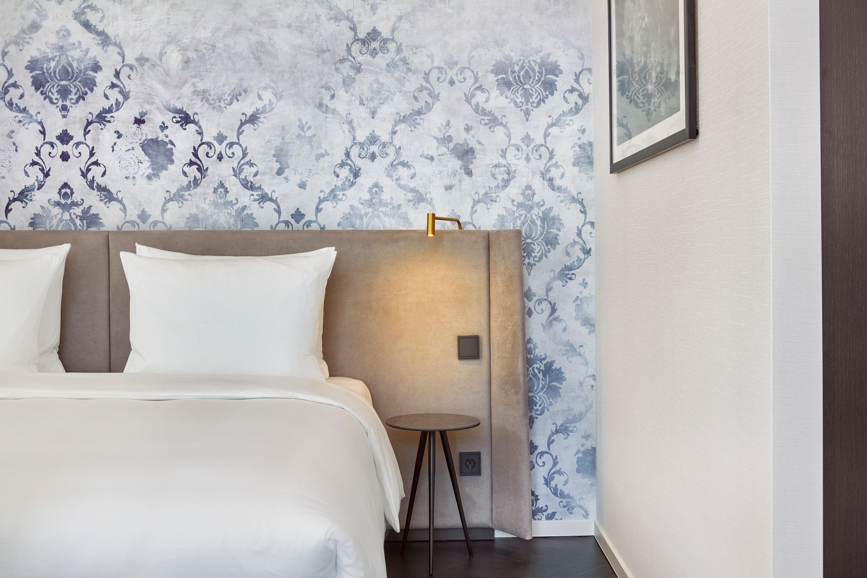 Modernes Hotelzimmer mit gemütlichem, beigem Polsterbett, weißen Bettwaren und elegantem Nachtisch. Dezente blaue Tapete mit floralem Muster schafft ruhige Atmosphäre. Wandlampe für stimmungsvolle Beleuchtung.