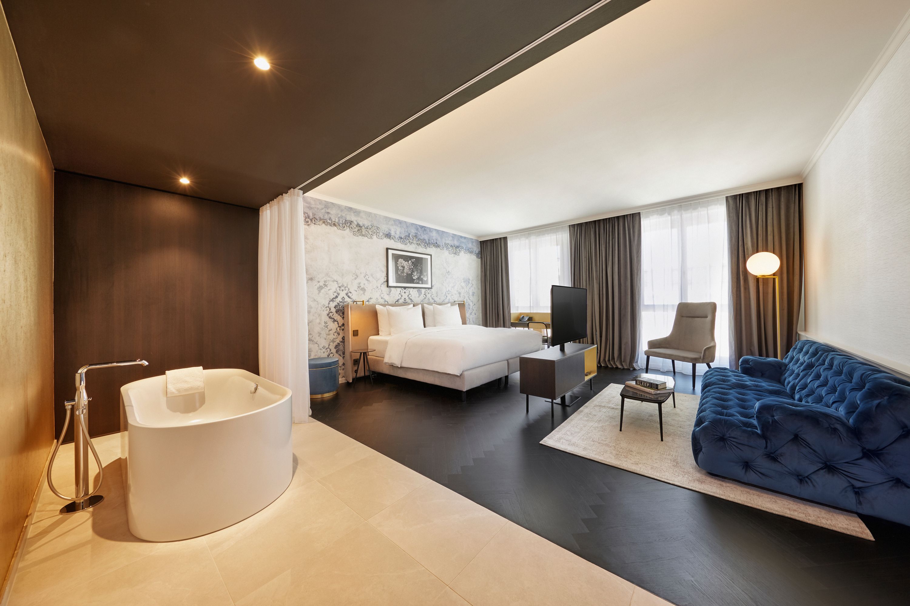 Modernes Hotelzimmer mit freistehender Badewanne, großem Doppelbett, gemütlicher Sitzecke mit dunklem Samtsofa und Stuhl, bodentiefen Fenstern und elegantem Ambiente.