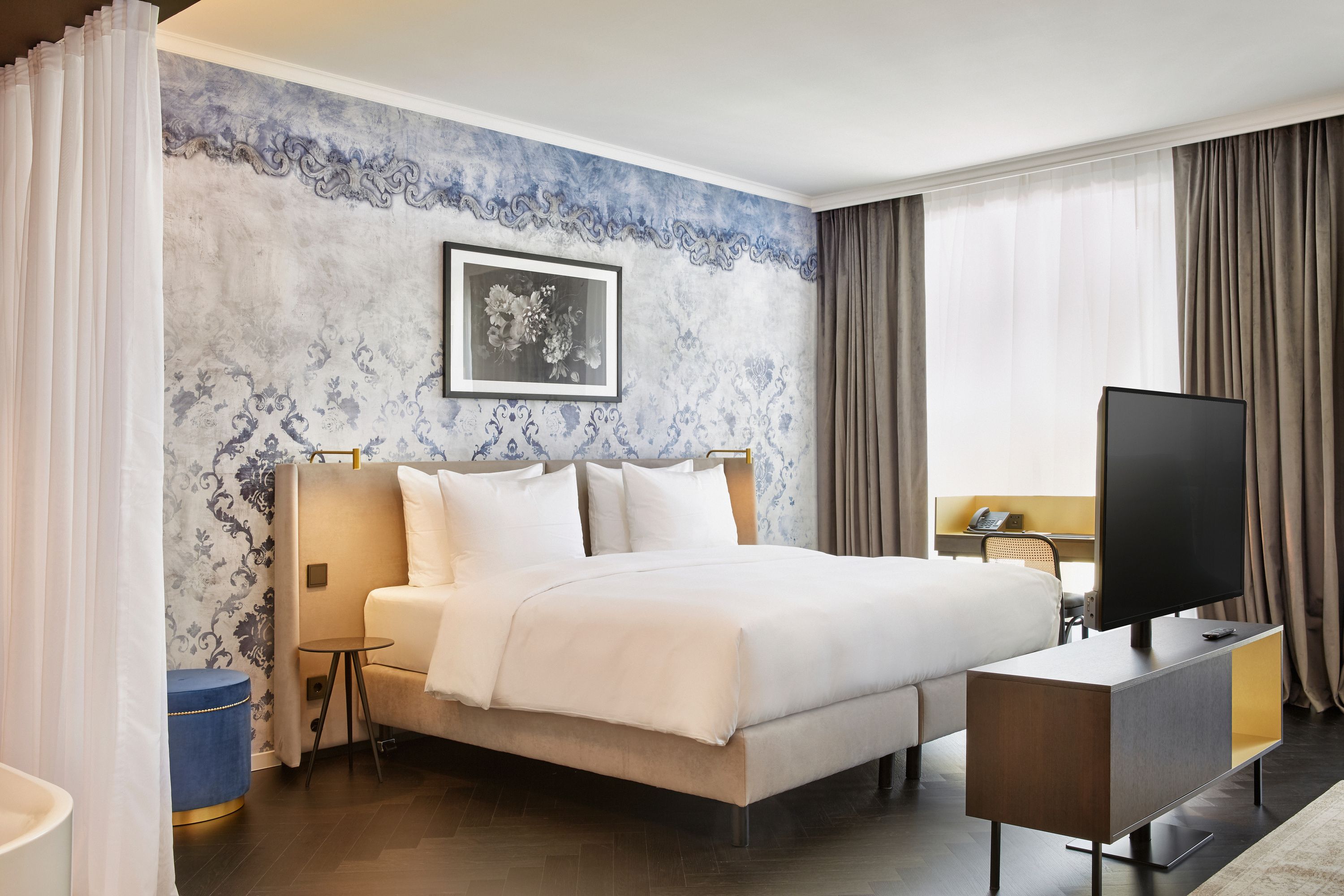 Modernes Hotelzimmer mit großem, weiß bezogenem Doppelbett, dekorativer blauer Wandgestaltung, großen Fenstern mit Vorhängen und einem Flachbildfernseher auf einem stilvollen Lowboard. Helle, elegante Atmosphäre für erholsamen Aufenthalt.