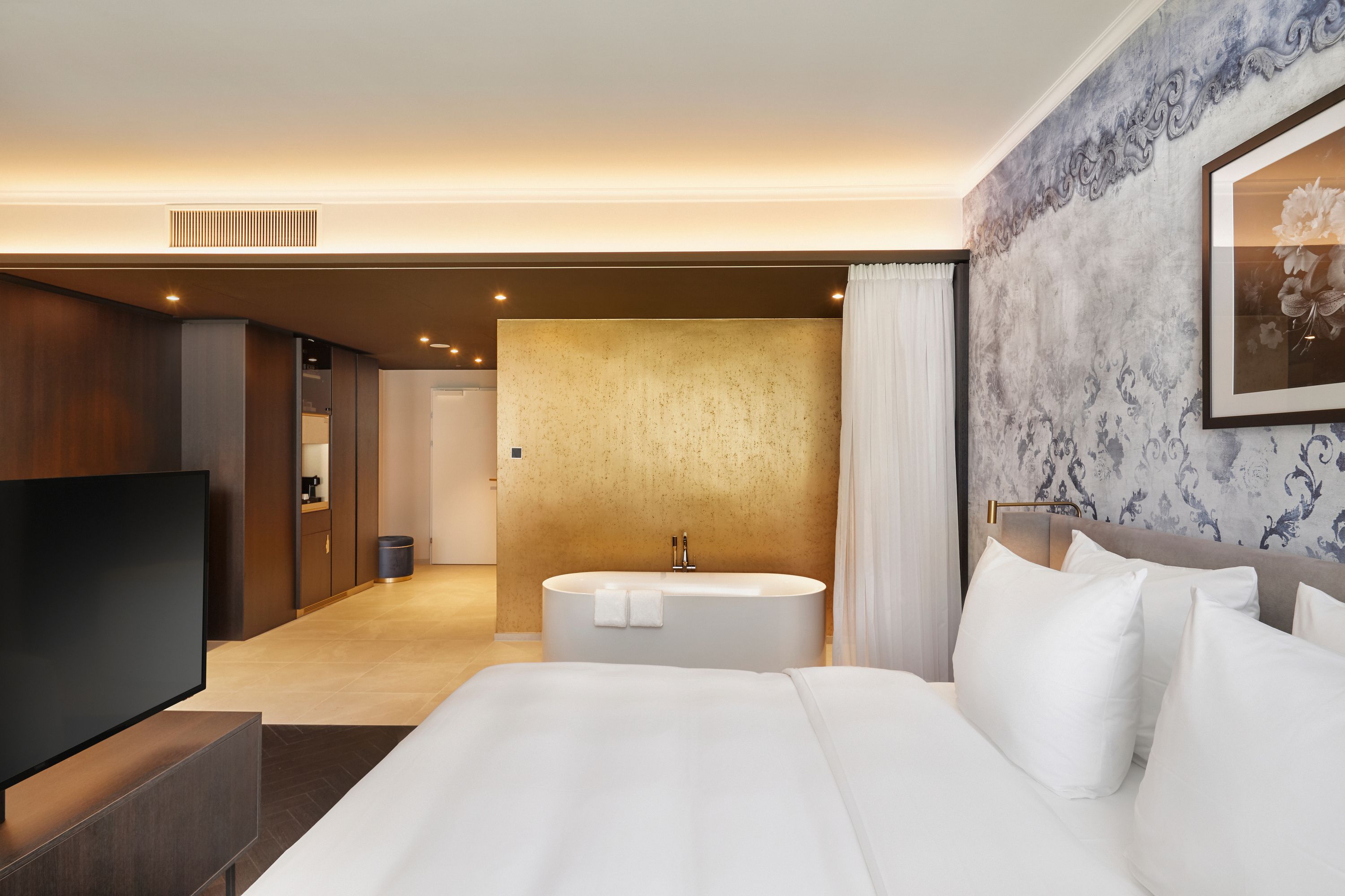 Modernes Hotelzimmer mit großem, weißem Bett, freistehender Badewanne vor goldfarbener Wand und dezent beleuchtetem Raum mit elegantem Wanddesign.