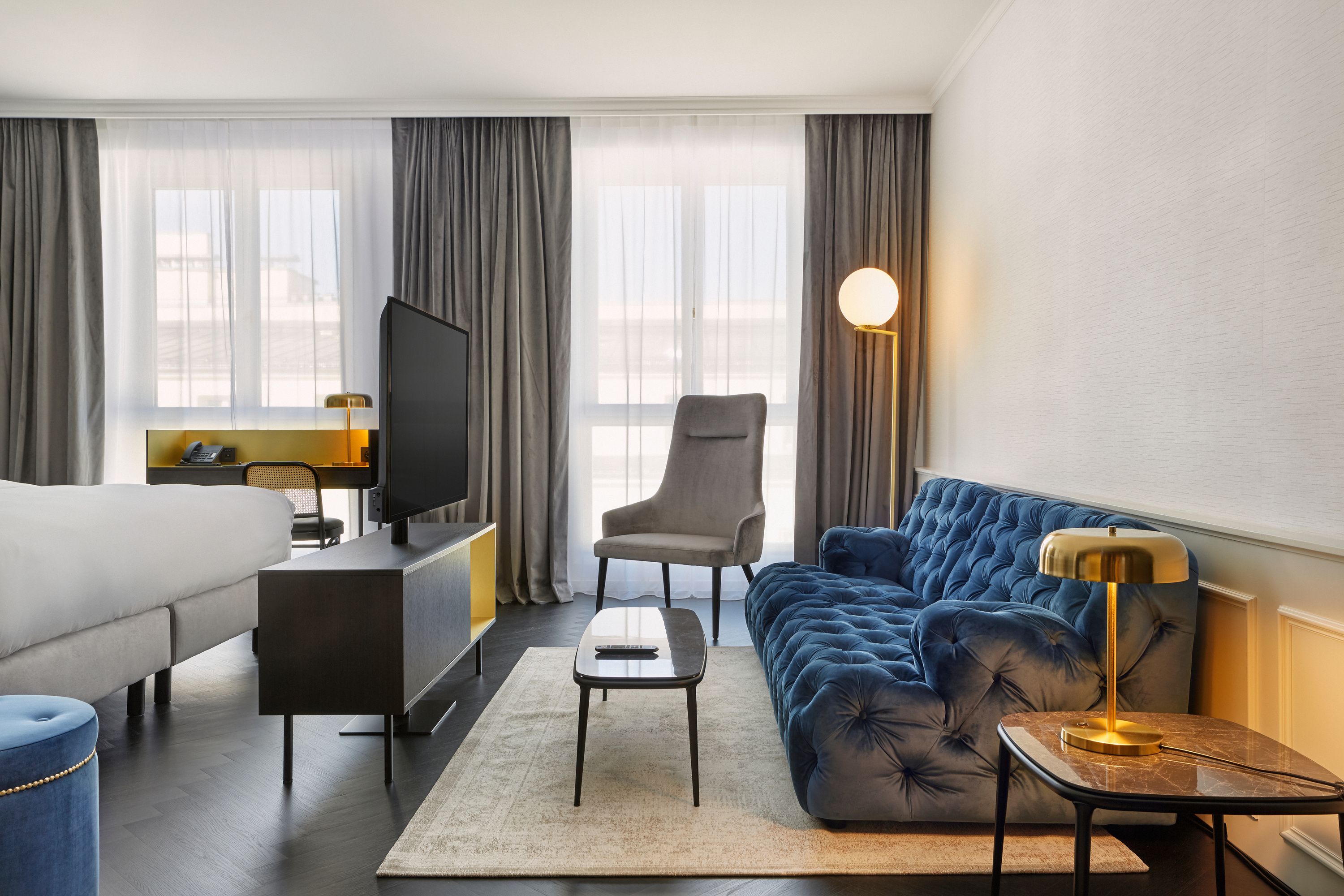 Modern eingerichtetes Hotelzimmer mit gemütlichem blauen Samtsofa, bequemer Sitzgelegenheit, Flachbildfernseher und großzügigem Tageslicht durch große Fenster mit halbtransparenten Vorhängen.