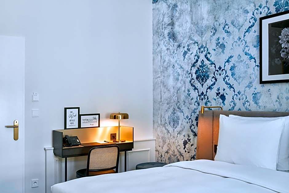 Elegantes Hotelzimmer mit großem, weiß bezogenem Bett, stilvoll gemusterten Wänden und einem kleinen Schreibtisch mit Lampe, Telefon und Stuhl, das eine ruhige Arbeits- und Entspannungsatmosphäre bietet.