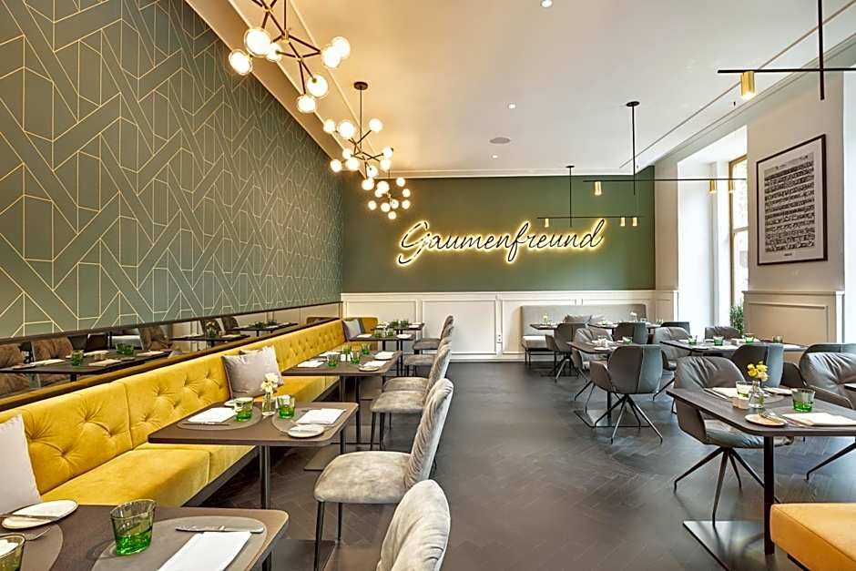 Modern eingerichtetes Restaurant mit gelber Polstersitzbank, grauen Stühlen, stilvollen Lampen und einem leuchtenden Schriftzug „Gaumenfreund“ an der Wand, das eine einladende Atmosphäre für genussvolle Mahlzeiten bietet.