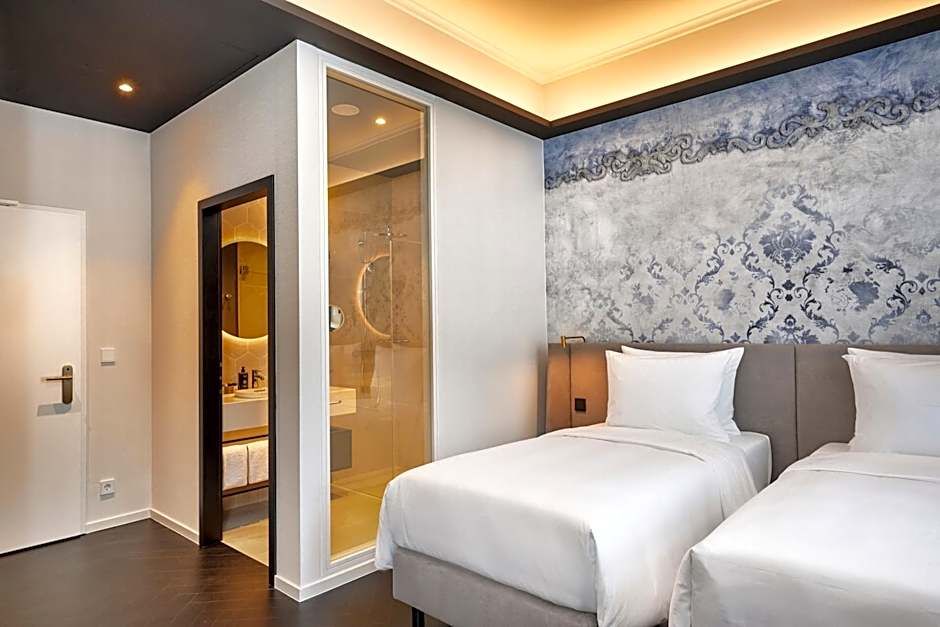 Modern eingerichtetes Hotelzimmer mit zwei Einzelbetten, stilvoller Wandgestaltung und indirekter Beleuchtung. Neben dem Zimmer befindet sich ein transparentes Badezimmer mit Dusche. Angenehme Atmosphäre für erholsamen Aufenthalt.