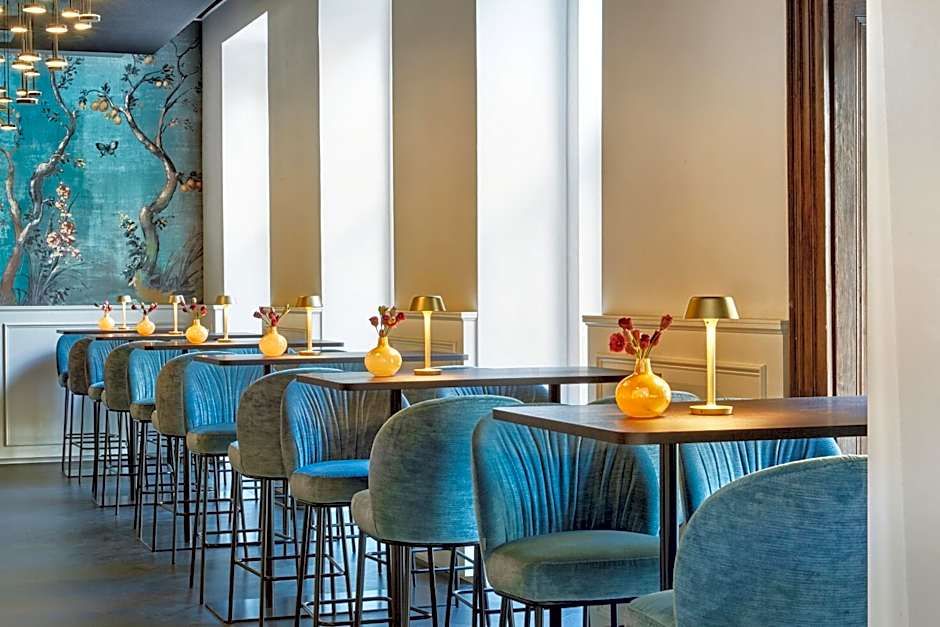 Elegantes Restaurant mit blauen Samtstühlen, Holztischen und moderner Dekoration. Natürliches Licht strömt durch große Fenster, eine beruhigende Wandgestaltung mit floralen Motiven sorgt für eine entspannte Atmosphäre. Kleine Vasen mit Blumen und stilvolle Tischlampen runden das Ambiente ab.