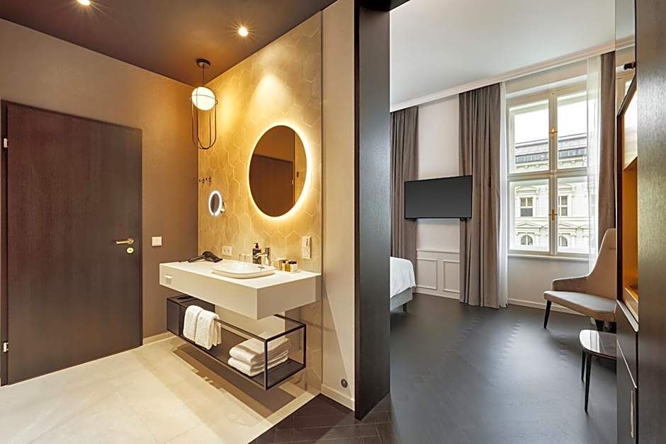 Modernes Badezimmer mit beleuchtetem runden Spiegel, Waschtisch und Handtüchern, angrenzend helles Schlafzimmer mit großem Fenster, Flachbildfernseher und komfortablem Sessel, stilvoll und einladend.