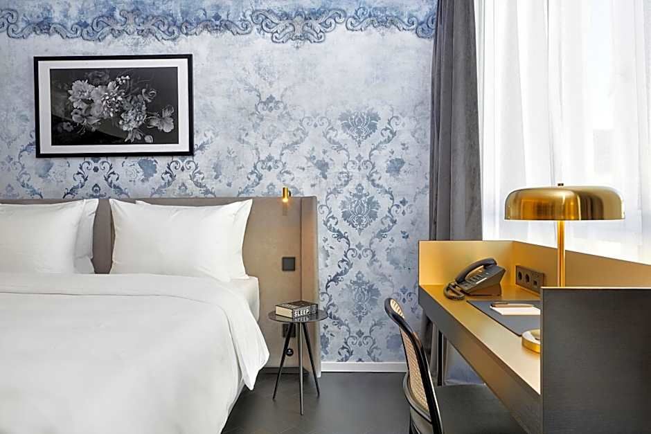 Hell eingerichtetes Hotelzimmer mit großem Bett, eleganter blauer Tapete, Schreibtisch mit moderner Lampe und Telefon, das Tageslicht durch leicht transparente Vorhänge hereinlässt.