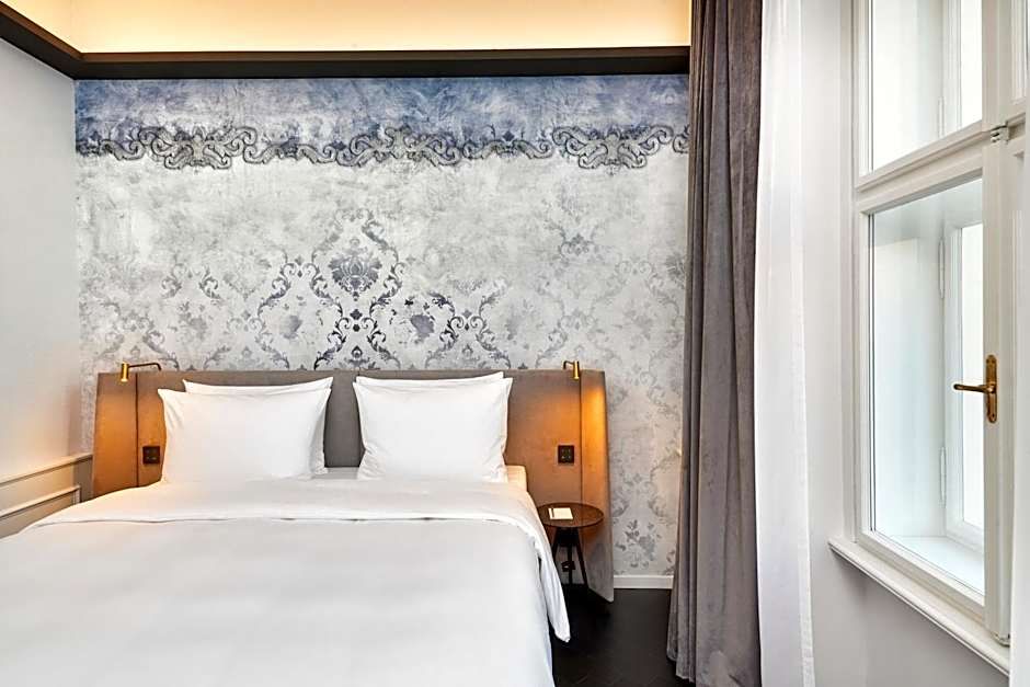 Modernes Hotelzimmer mit großem Doppelbett, weißer Bettwäsche und dekorativer, stilvoller Tapete. Natürliches Licht fällt durch das Fenster, das mit Vorhängen ausgestattet ist, und daneben befinden sich zwei kleine Nachttische mit Leselampen.