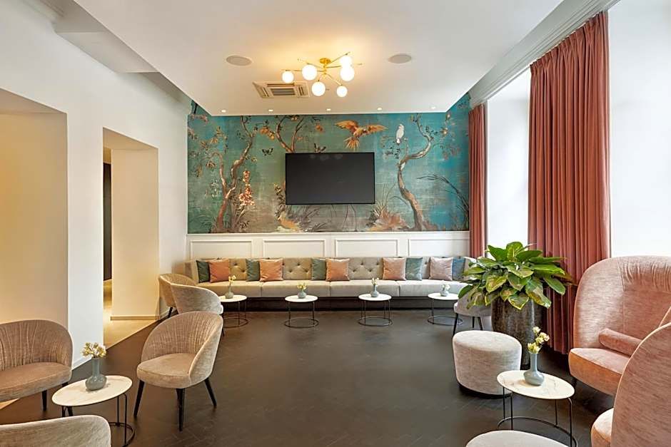 Helles, modernes Lounge-Zimmer mit langer Couch, weichen Sesseln und runden Tischen. Eine Wand zeigt kunstvolle Naturmotive, große Fenster mit roten Vorhängen lassen Tageslicht herein. Ruhige Atmosphäre zum Entspannen oder für Gespräche.