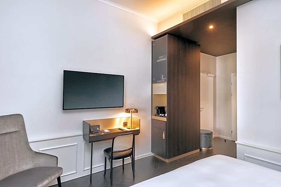 Helles Hotelzimmer mit modernem Schreibtisch, Stuhl, Wand-TV und gemütlicher Beleuchtung, daneben ein grauer Sessel und ein offener Schrank mit Kaffeezubehör.