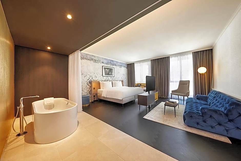 Modernes Hotelzimmer mit offenem Grundriss, Badewanne im Raum, großem Doppelbett, samtblauem Sofa, einem Sessel und bodentiefen Fenstern, die viel Tageslicht hereinlassen.