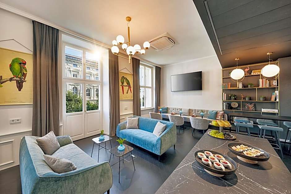 Modern eingerichteter Aufenthaltsraum mit gemütlichen Sofas, großem TV, dekorativen Papageien-Motiven an der Wand und einem Buffet mit Snacks. Helle Fenster bieten Tageslicht und Blick auf benachbarte Gebäude. Ideal zum Entspannen und geselligen Beisammensein.