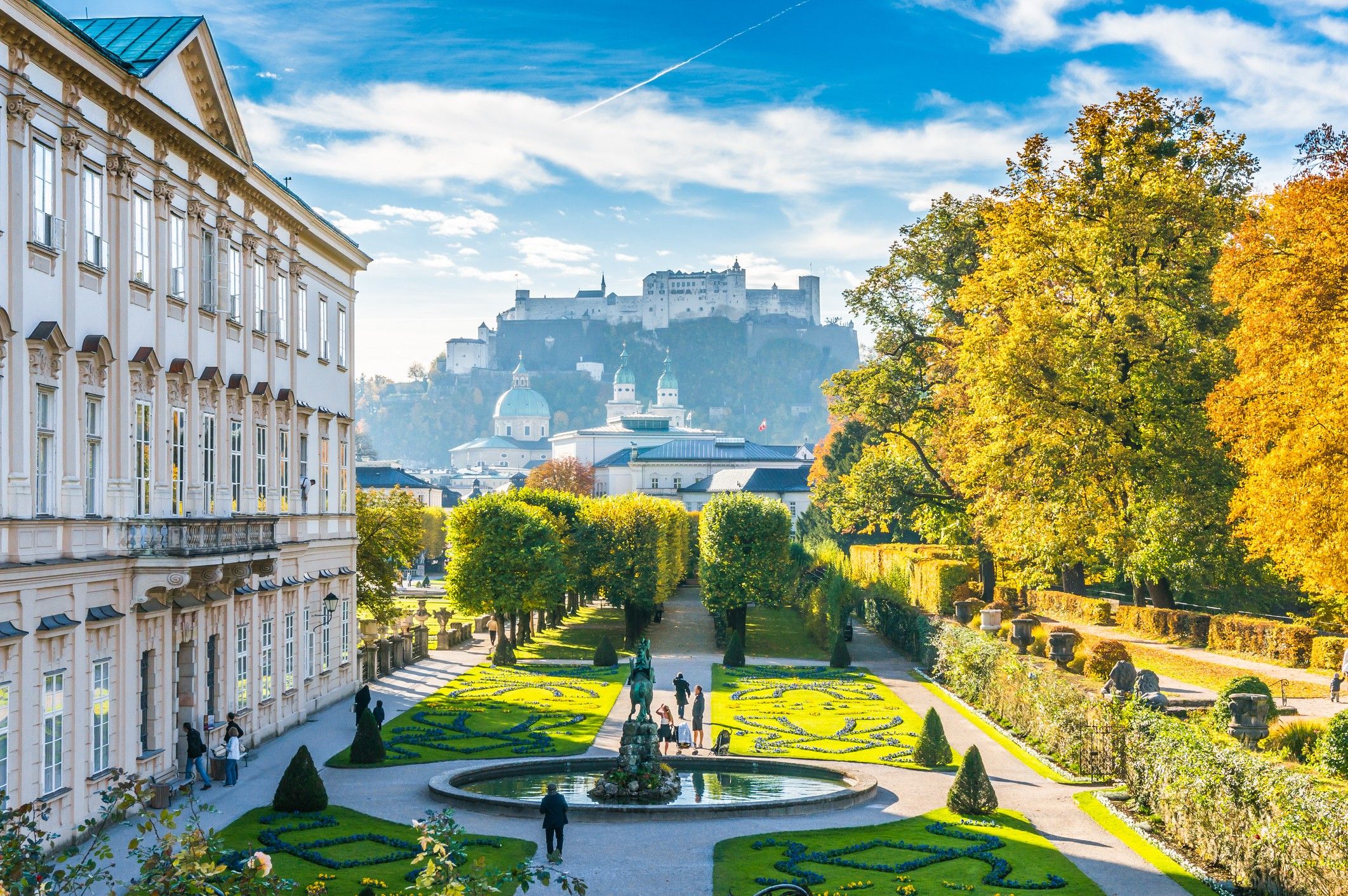 Blick auf einen gepflegten Barockgarten mit kunstvoll geschnittenen Hecken und Springbrunnen, umgeben von historischen Gebäuden, mit der Festung Hohensalzburg im Hintergrund unter blauem Himmel.