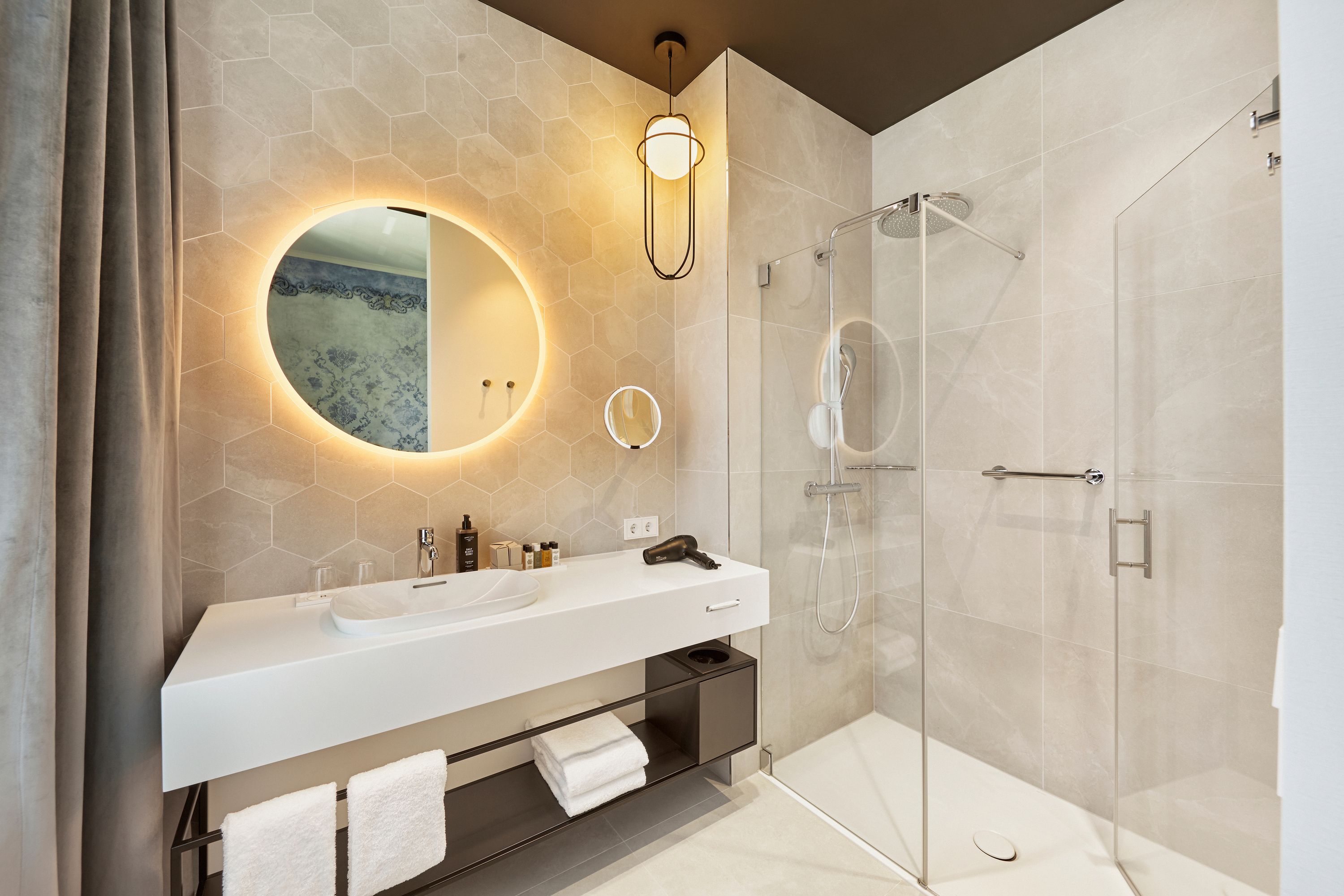 Modernes, helles Badezimmer mit großer begehbarer Dusche, rundem, beleuchtetem Spiegel und weißem Waschtisch. Nützliche Accessoires wie Handtücher, Föhn und Kosmetikspiegel sorgen für Komfort. Klare, schlichte Gestaltung schafft eine elegante und einladende Atmosphäre.