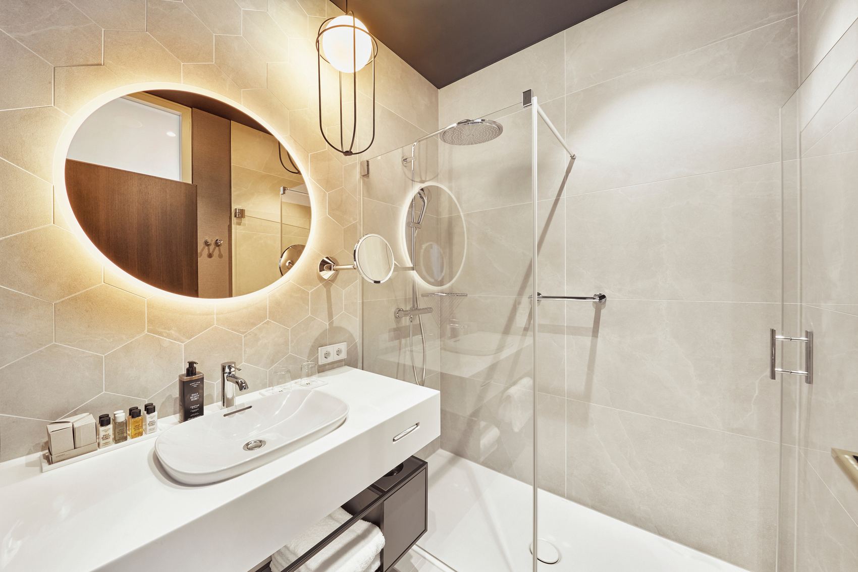 Modernes Badezimmer mit beleuchtetem Rundspiegel, Waschtisch mit Waschbecken und Pflegeprodukten sowie großzügiger, ebenerdiger Dusche mit Glasabtrennung. Helle, elegante Fliesen und stilvolle Beleuchtung schaffen eine angenehme Atmosphäre.