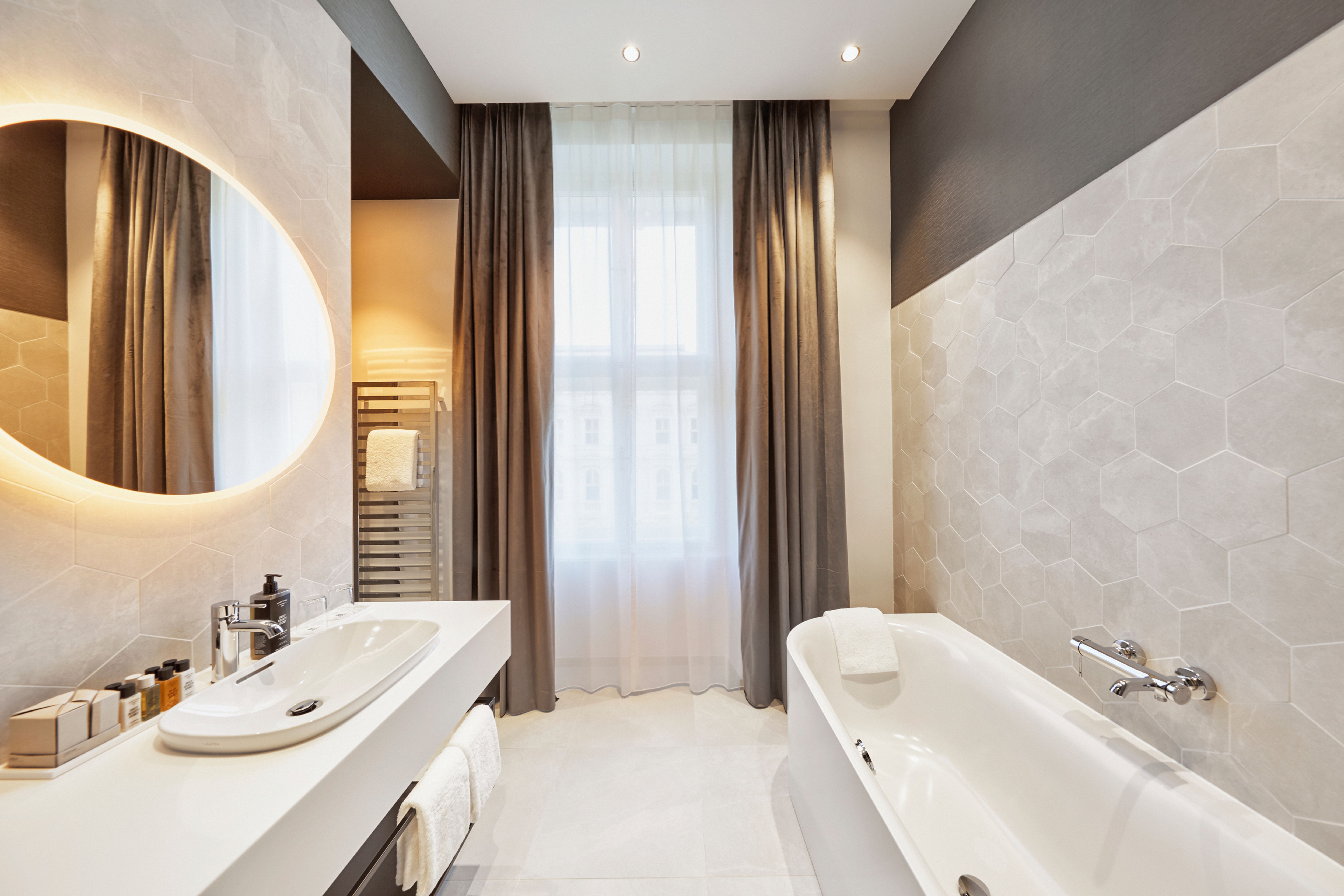 Modernes, helles Badezimmer mit freistehender Badewanne, großem beleuchtetem Spiegel, elegantem Waschbecken und großen Fenstern mit Vorhängen, das eine entspannende und komfortable Atmosphäre vermittelt. Modernes, helles Badezimmer mit freistehender Badewanne, großem beleuchtetem Spiegel, elegantem Waschbecken und großen Fenstern mit Vorhängen, das eine entspannende und komfortable Atmosphäre vermittelt.